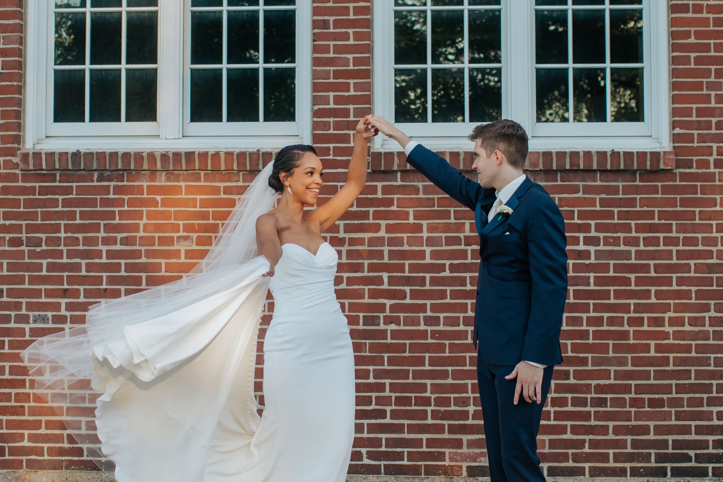 A wedding party stands in front of a red brick building with white trim windows during a formal photo session.