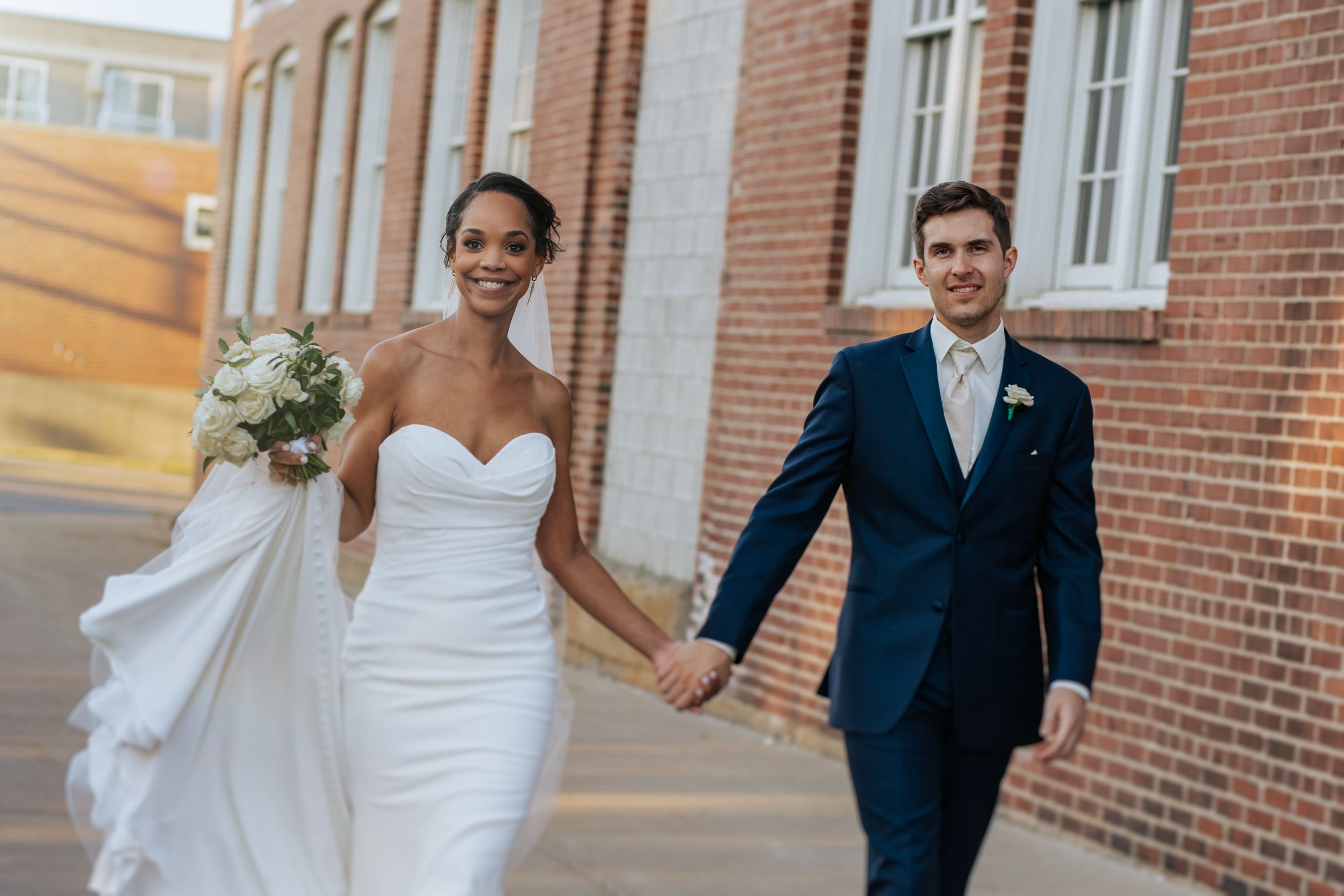 A couple in wedding attire runs playfully along a brick building exterior during golden hour.