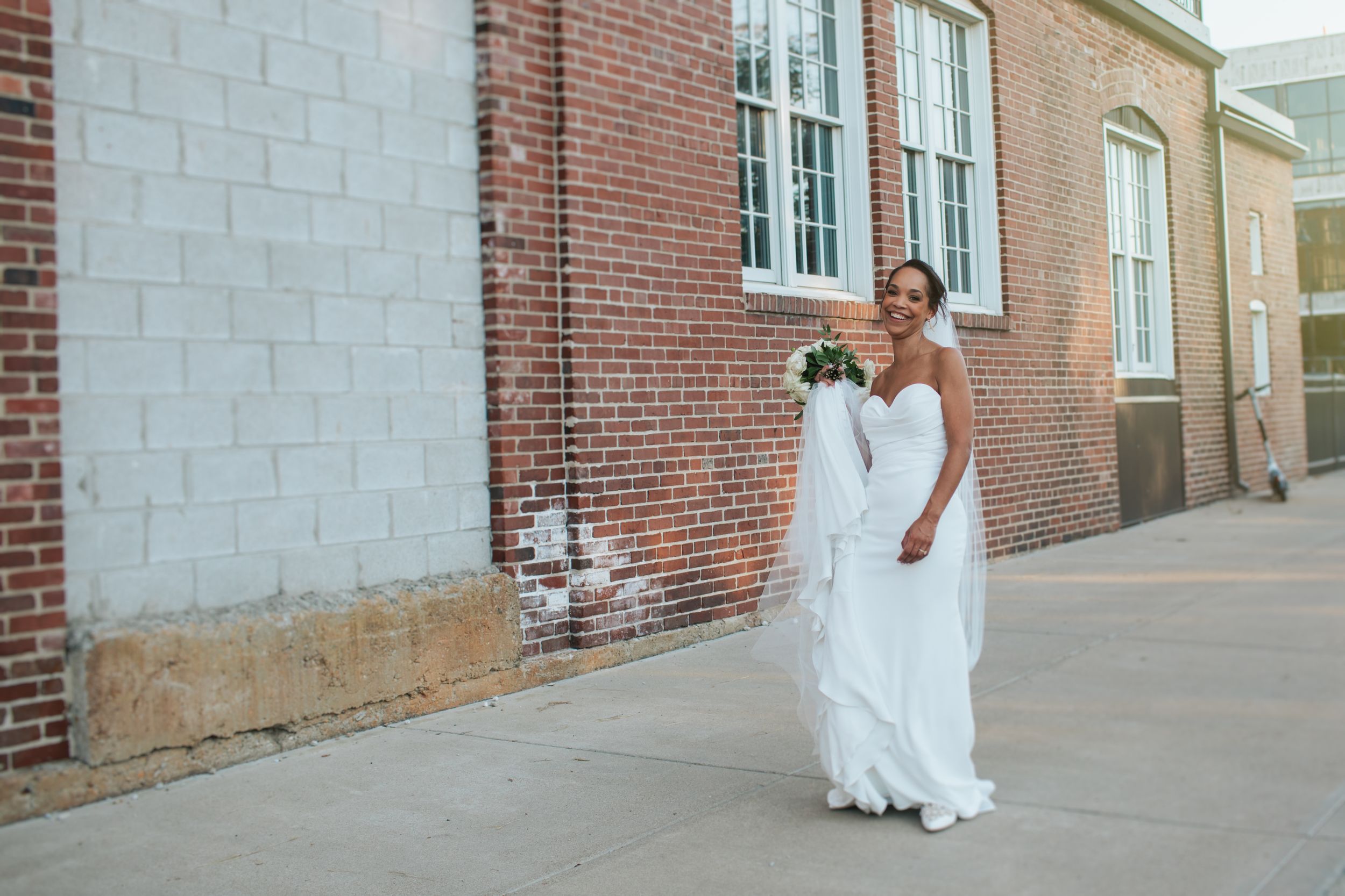 A person in a flowing white dress walks along a brick wall in an urban alleyway.