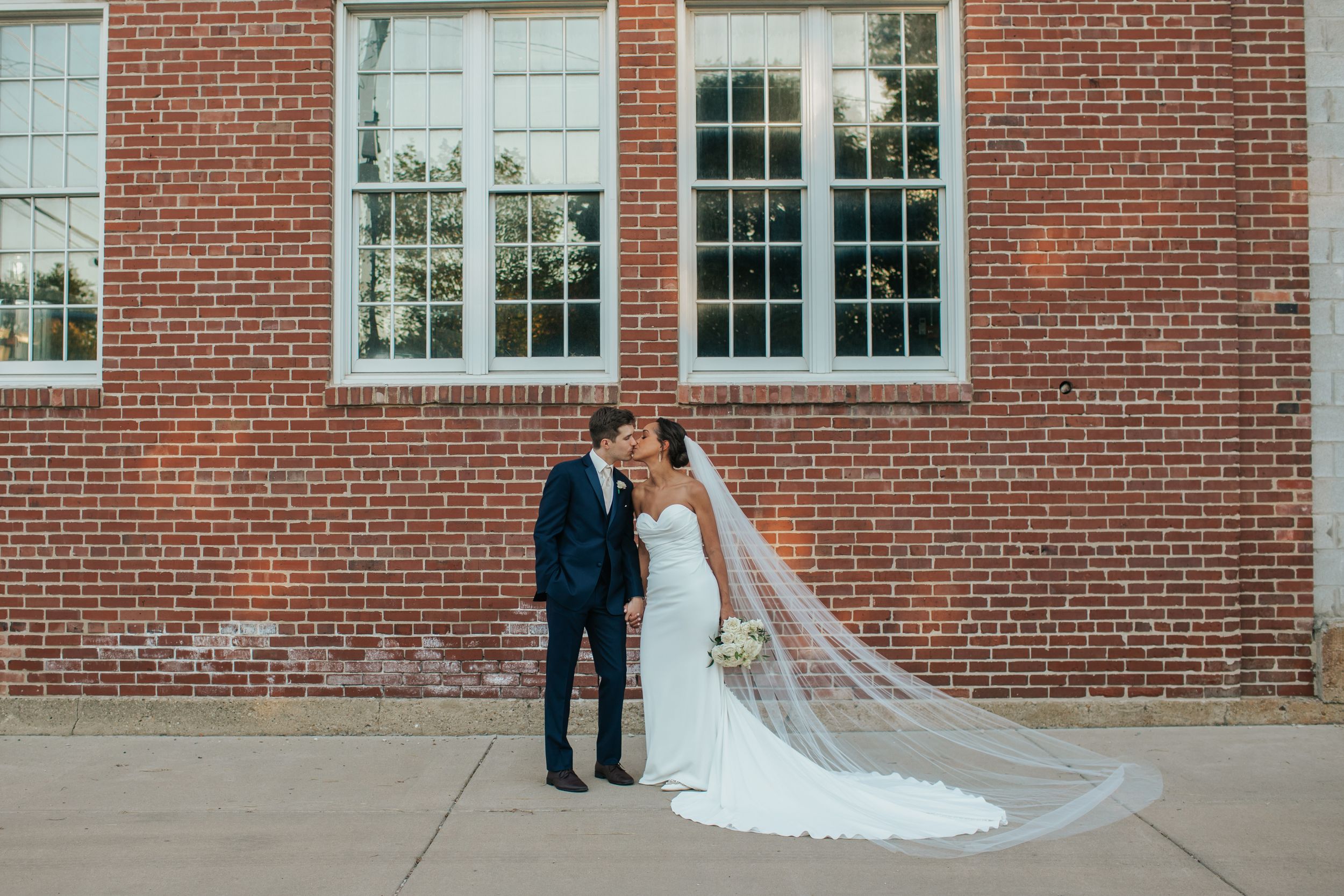 A couple shares a romantic kiss outside a historic brick building with white trim windows and a flowing wedding veil.