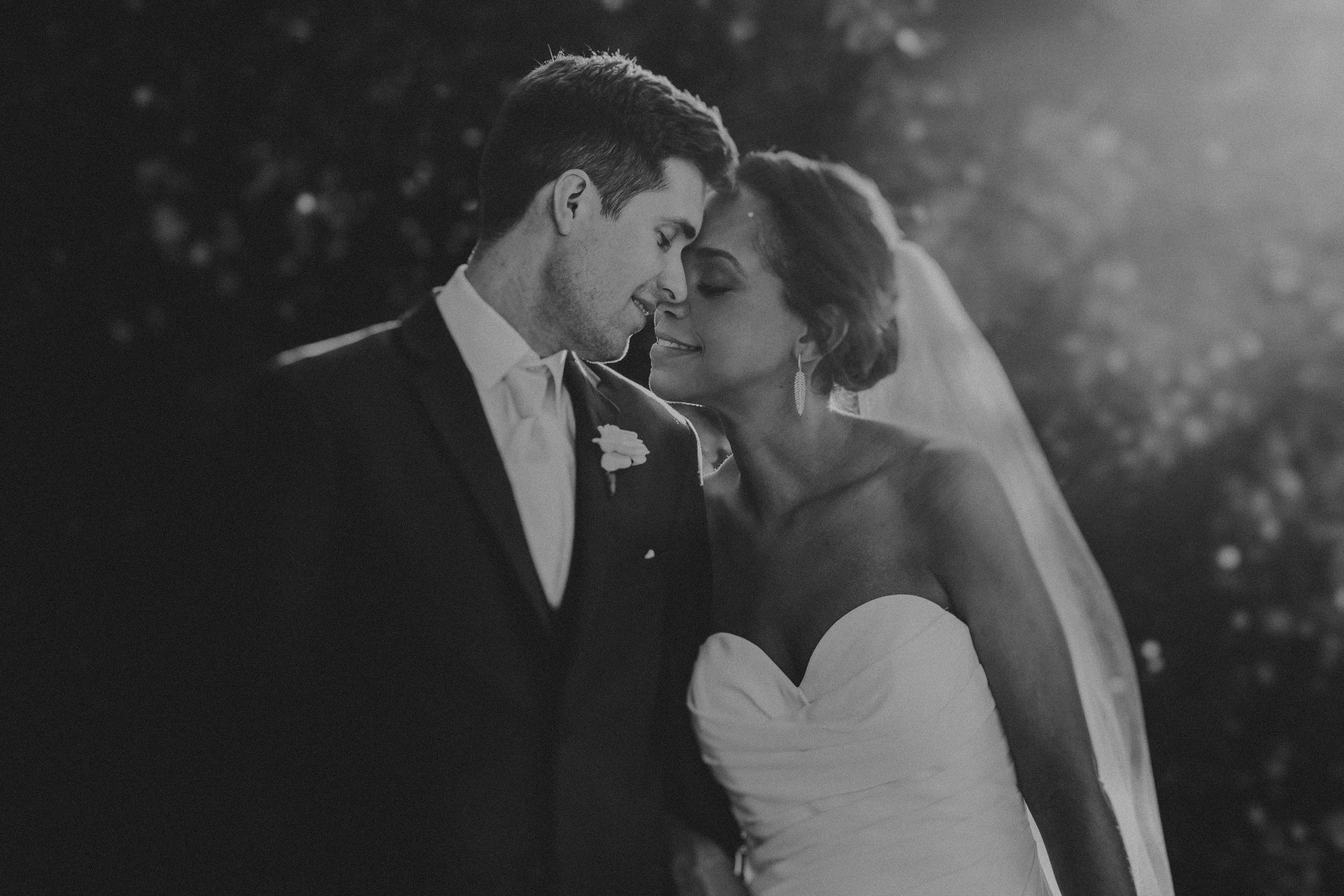 A romantic black and white wedding photo of a couple in formal attire sharing an intimate moment outdoors.