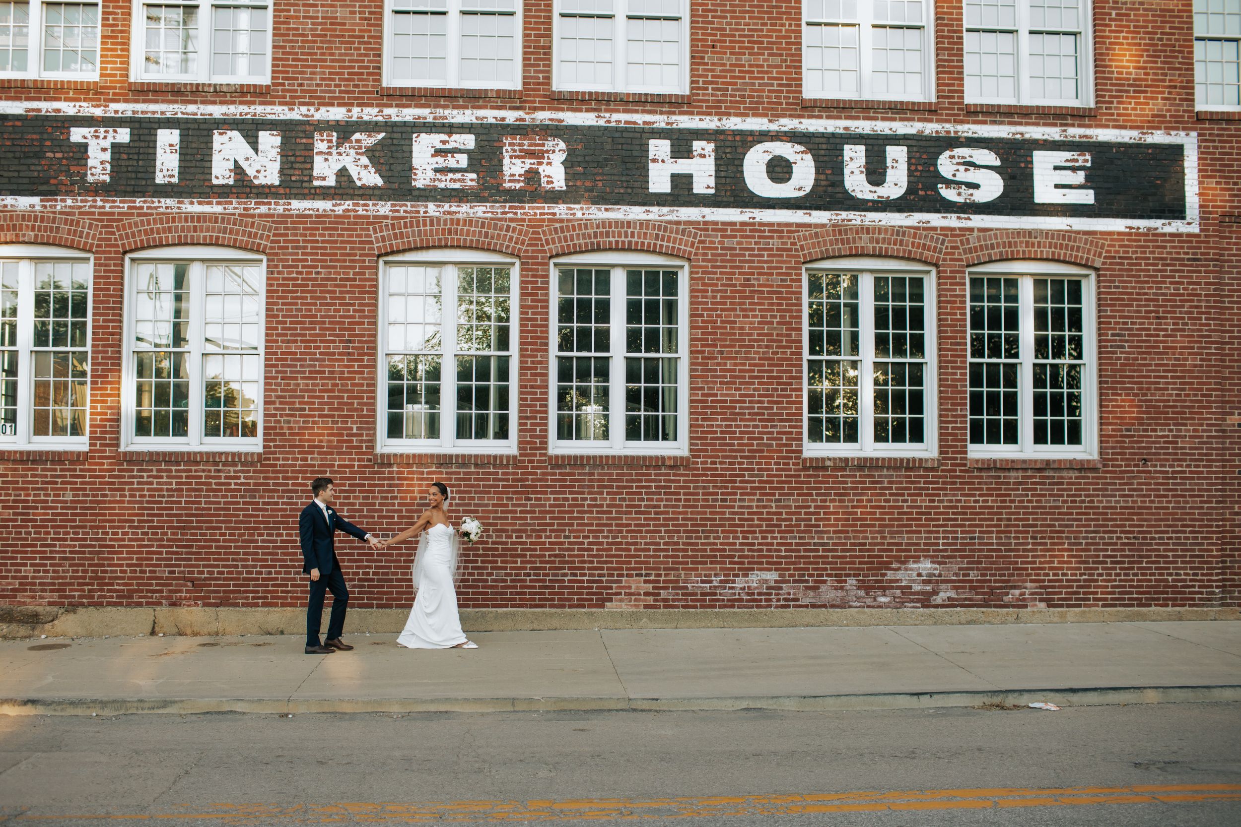 A couple stands outside the brick facade of Tinker House building with large white-framed windows.