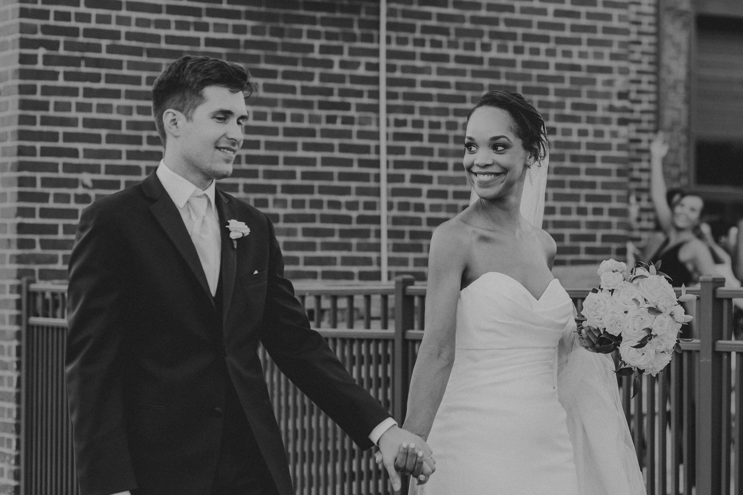 Wedding party members walk and laugh together on a brick patio during an outdoor celebration.