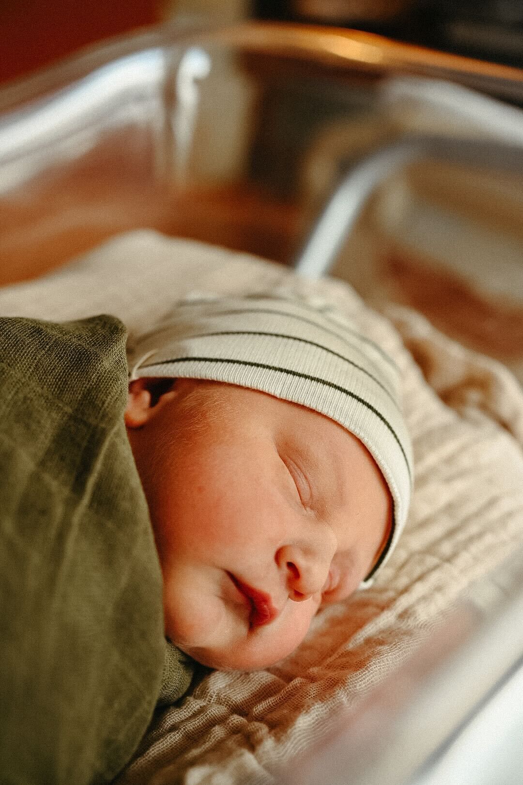 Sleeping newborn wearing beige hat in hospital bassinet.
