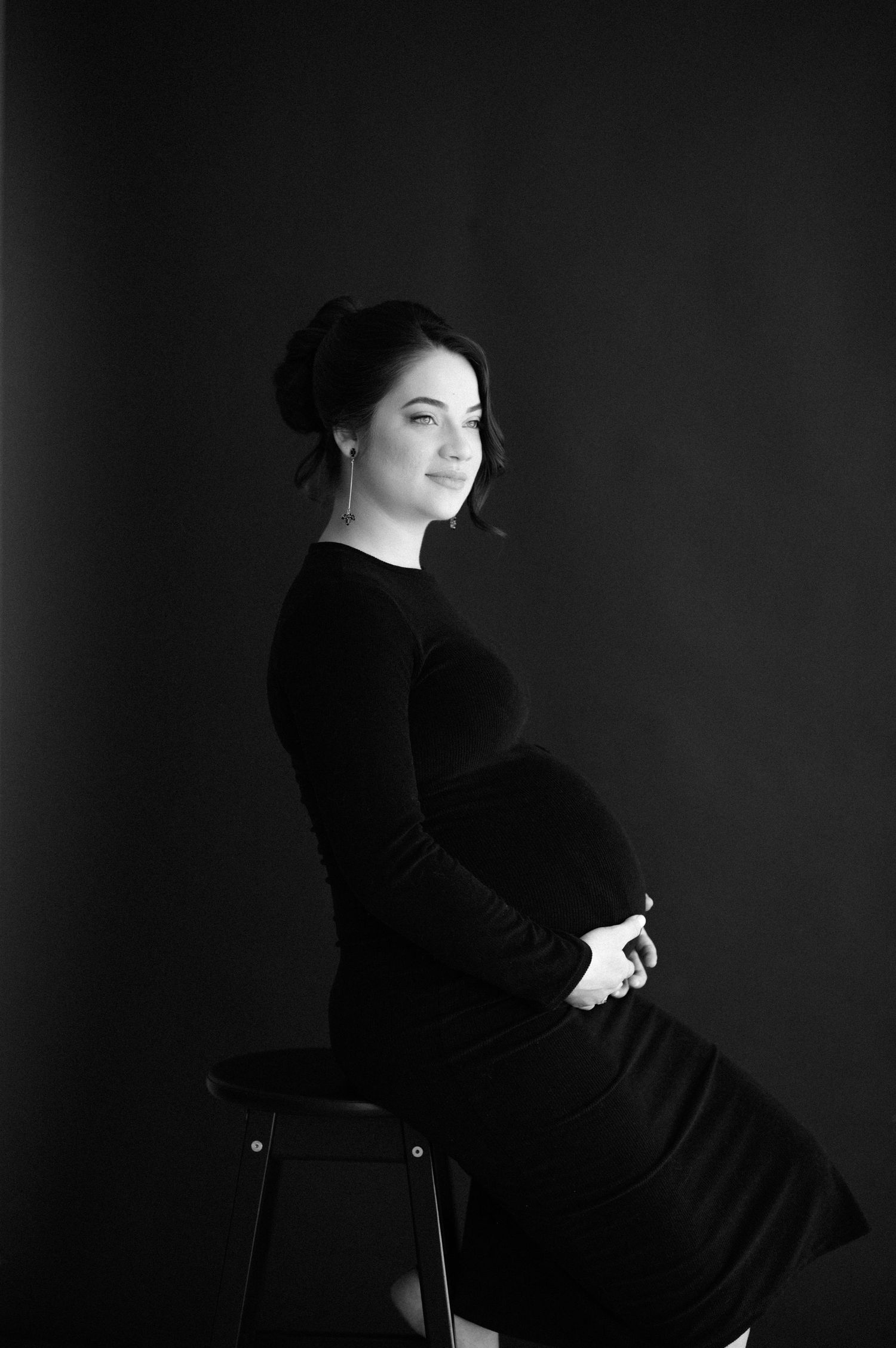 Black and white maternity portrait of woman in dark dress seated elegantly against dark background.