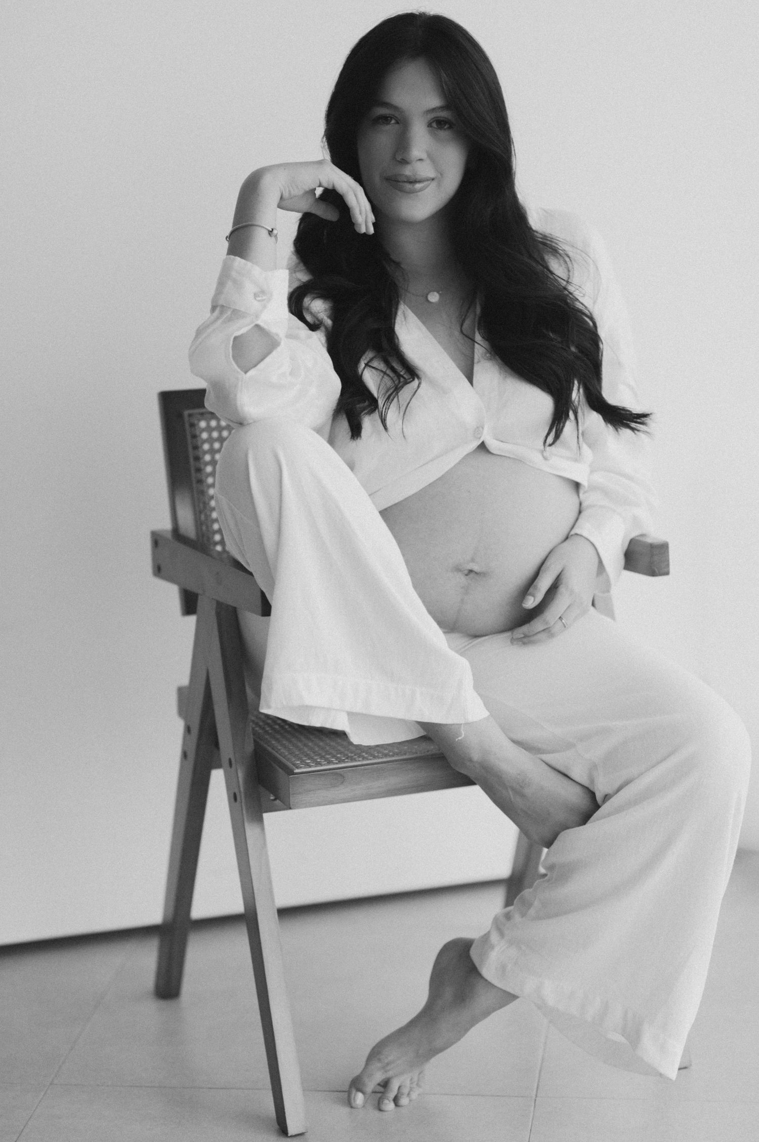 Black and white maternity photo of a woman in white crop top and pants sitting on a chair cradling her baby bump.
