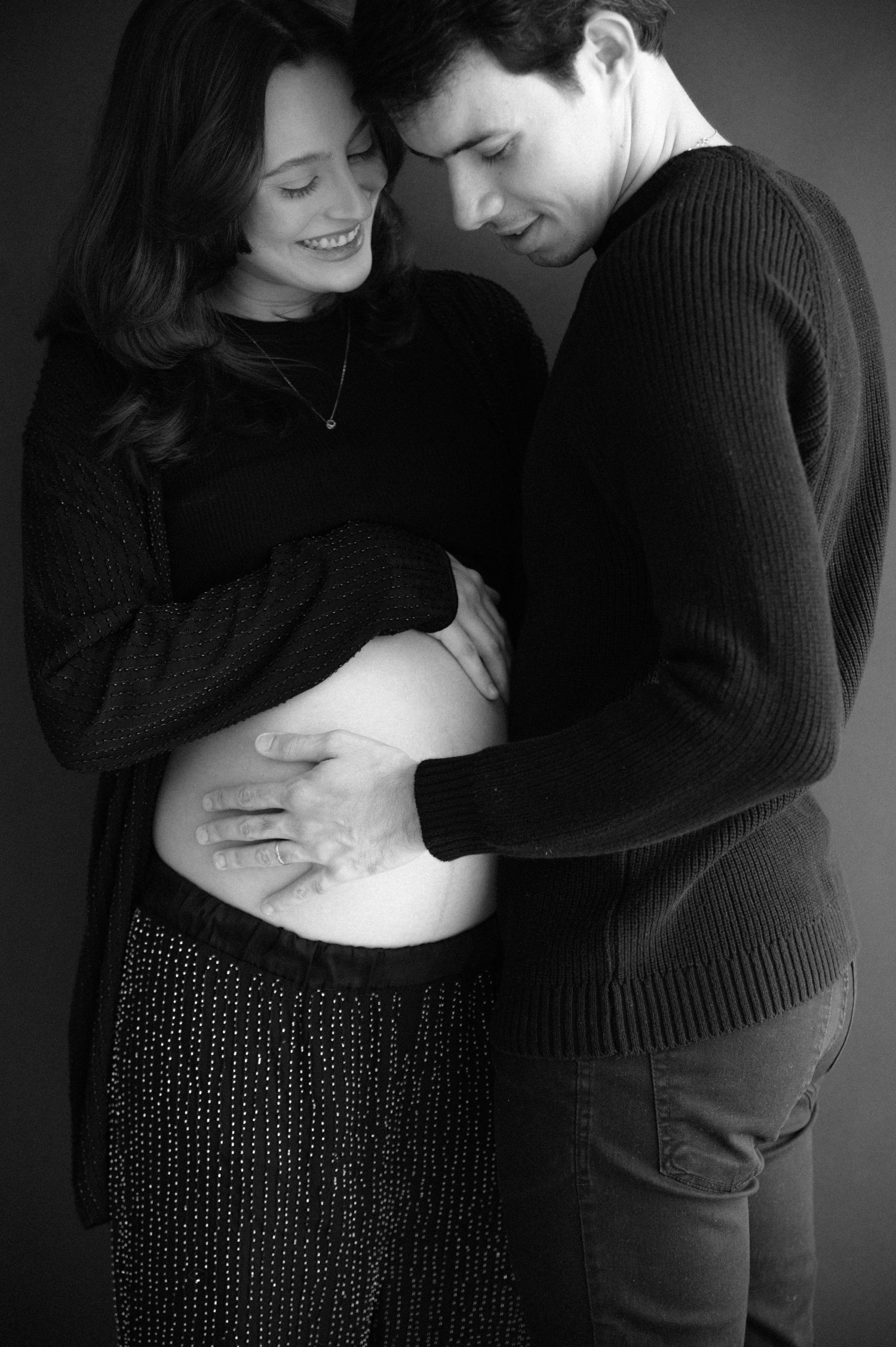 A black and white maternity photo shows an intimate moment between a couple as they tenderly touch her pregnant belly.