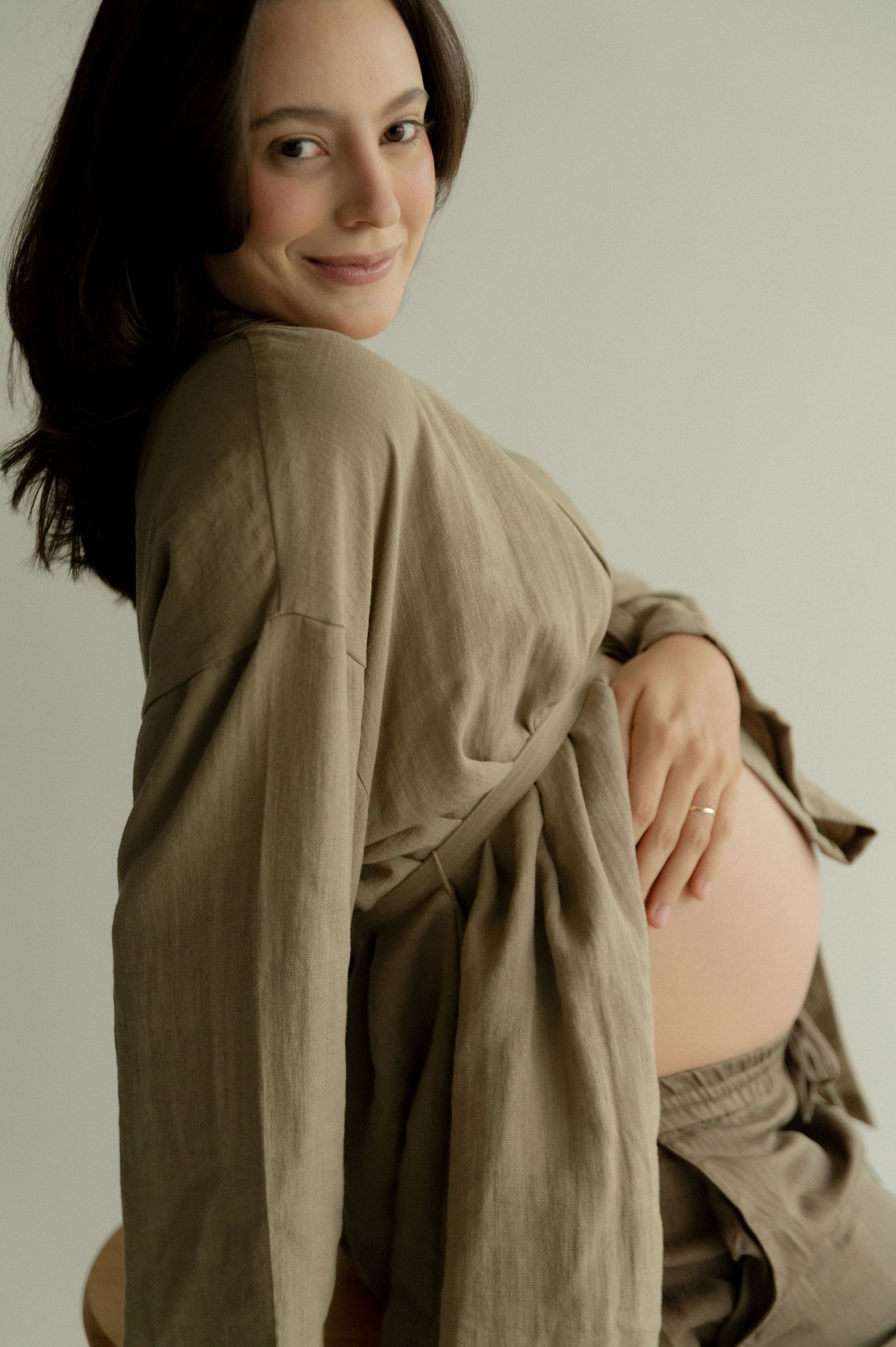 A pregnant woman in a beige linen dress poses sideways against a neutral background in a minimalist maternity photo.