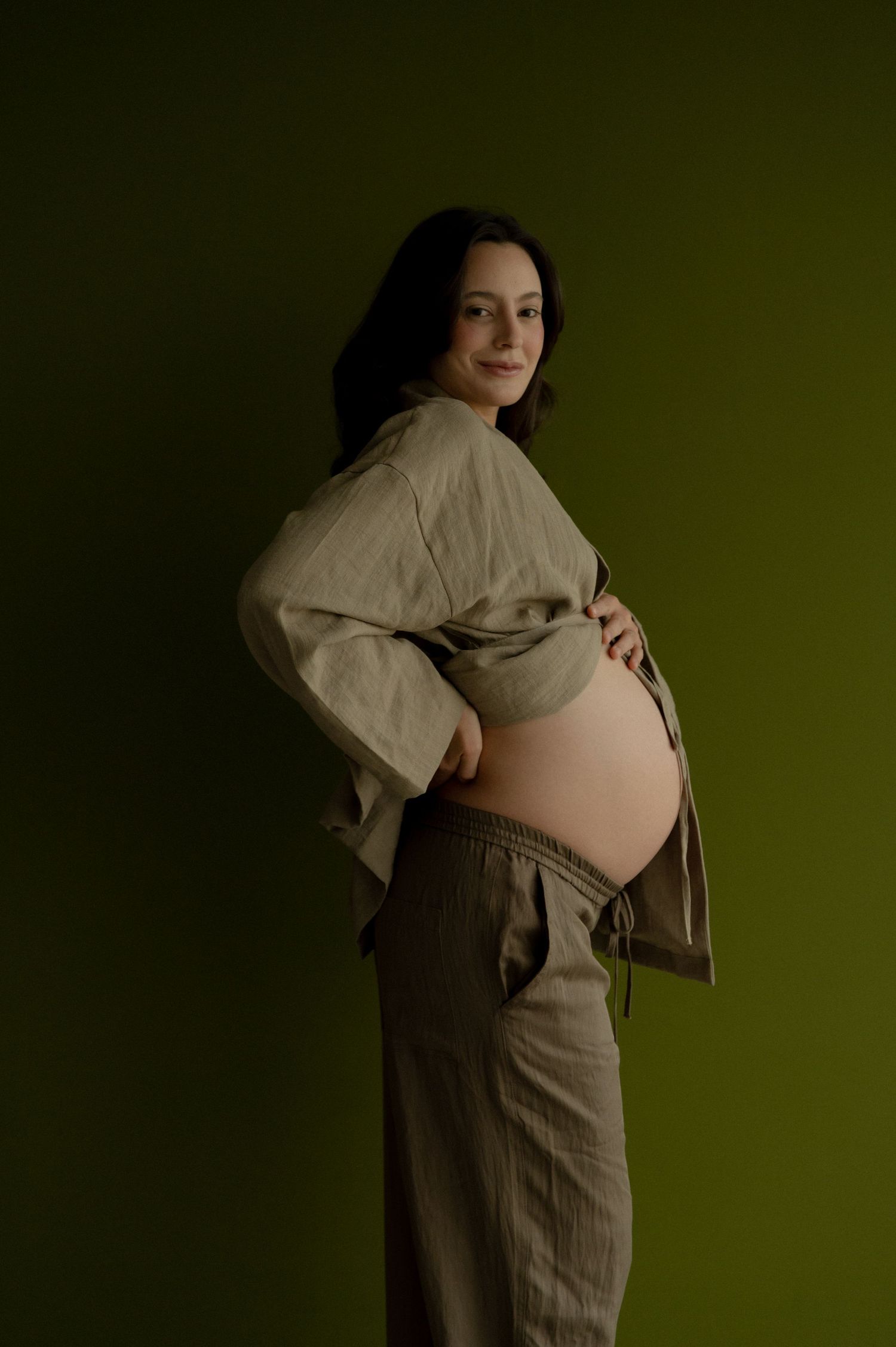 A pregnant person in casual linen clothing poses against a dark green background in a maternity photoshoot.