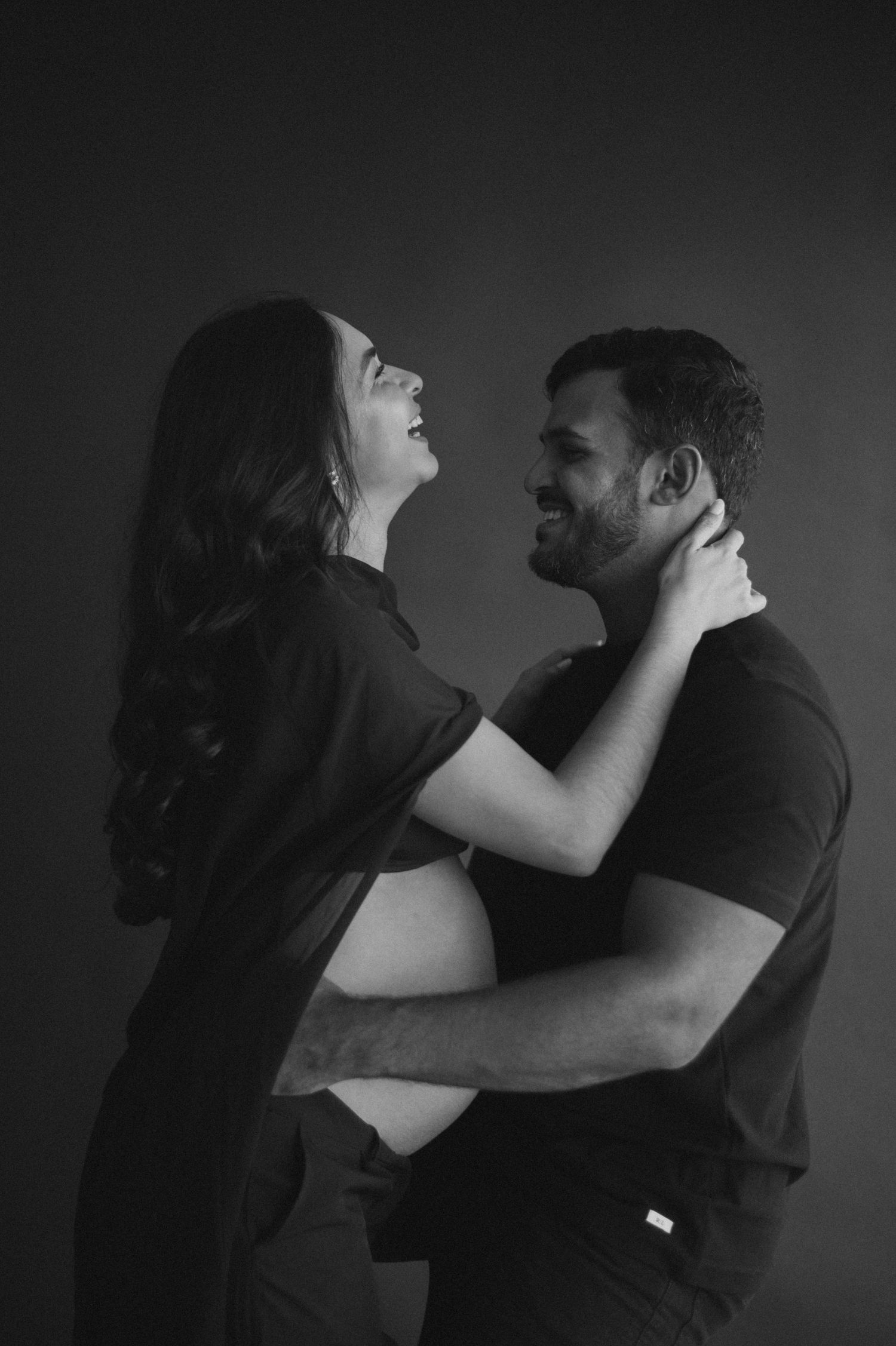 Black and white maternity photo of a smiling couple embracing, the woman showing her baby bump, against a dark background.