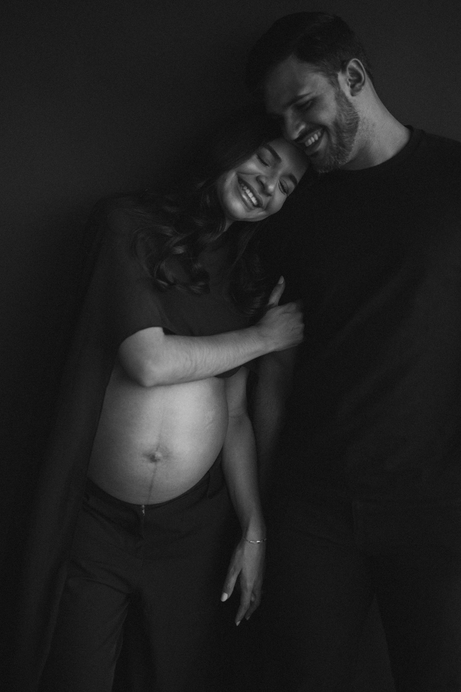 Black and white maternity photo of a smiling couple embracing, showing pregnant belly, studio portrait.