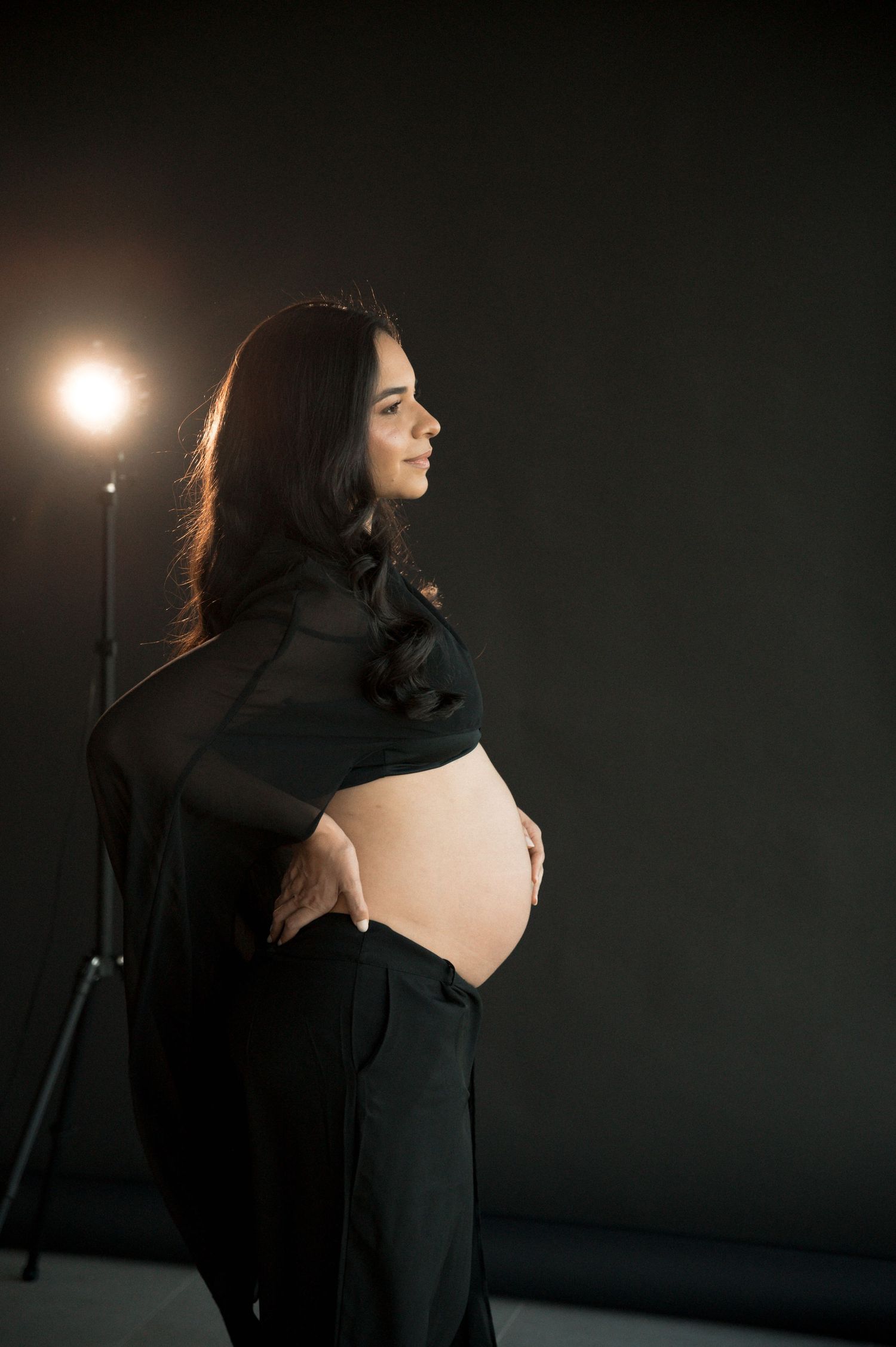 Pregnant woman in black outfit cradling bare baby bump in dark studio with dramatic lighting.