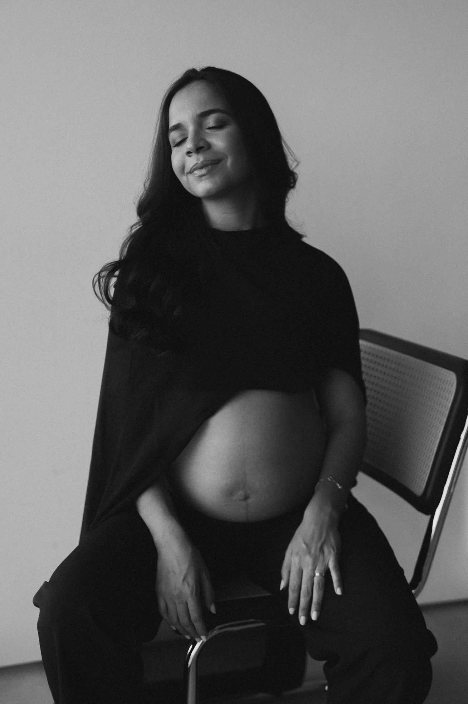Black and white maternity photo of a serene pregnant woman sitting in a chair, eyes closed, cradling her bare baby bump.