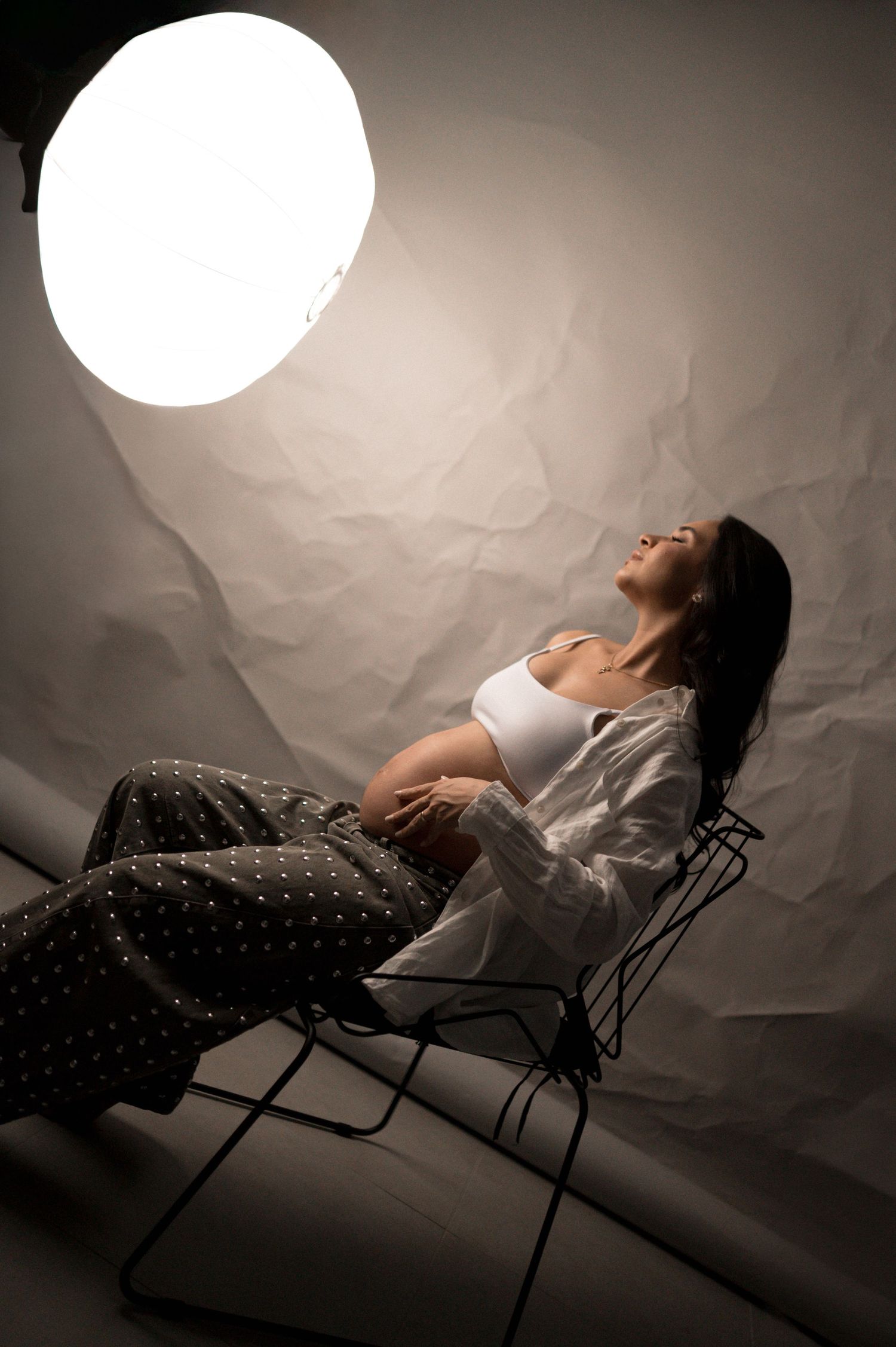 Pregnant woman reclines on chair under studio light, cradling belly, wearing white top and polka dot pants.