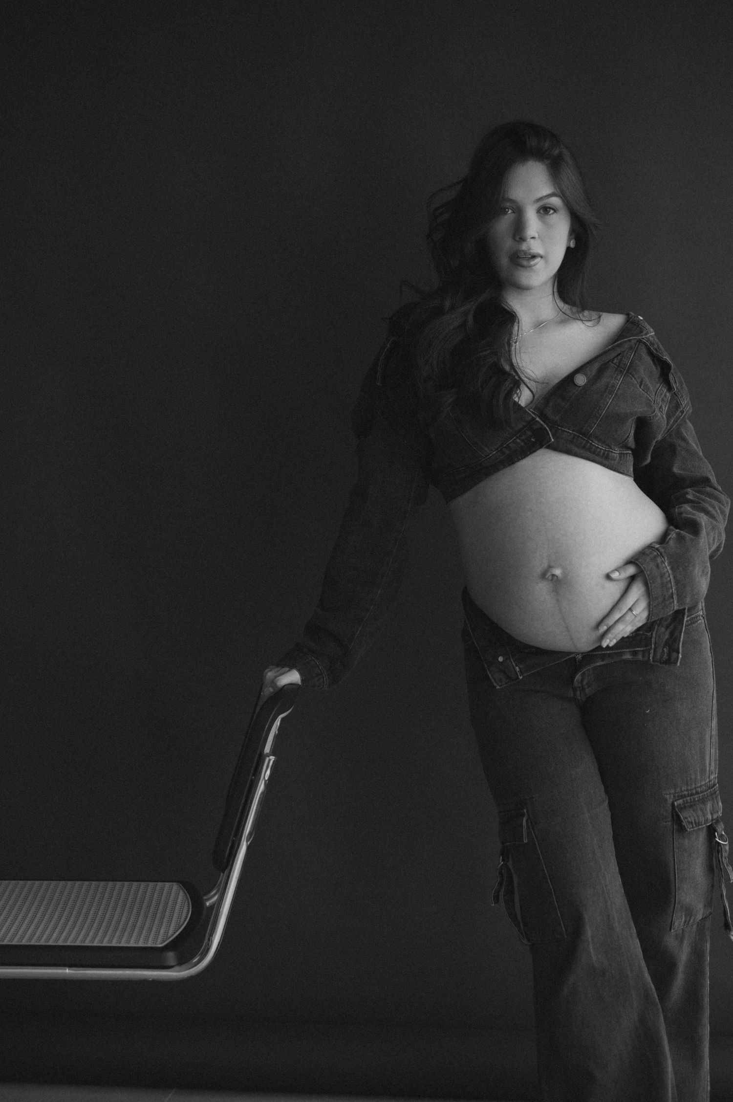 Black and white maternity photo of a woman in denim jacket and jeans posing with a chair against dark background.