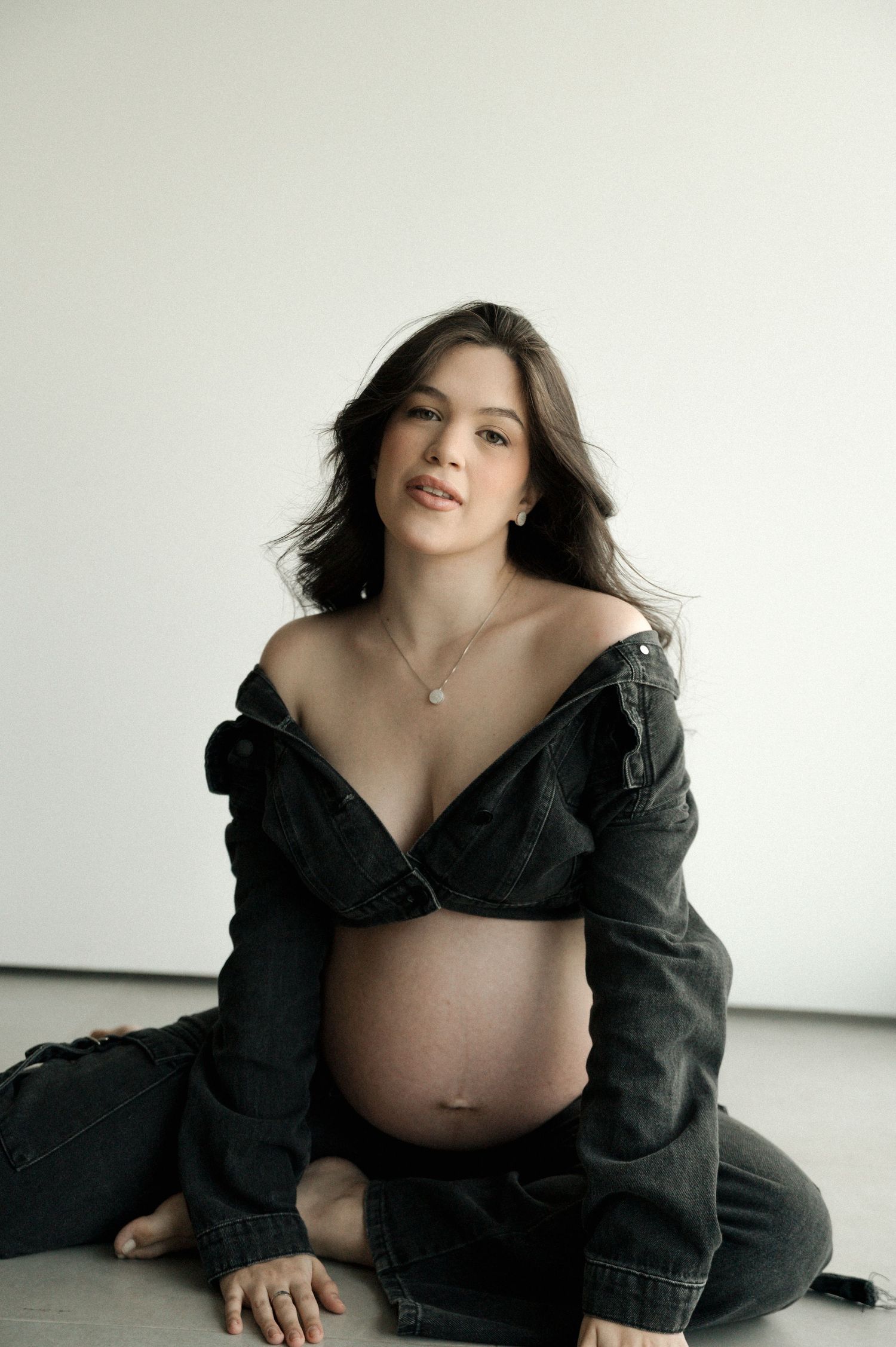 Pregnant woman in black denim jacket sitting on floor, showcasing baby bump in a minimalist studio setting.