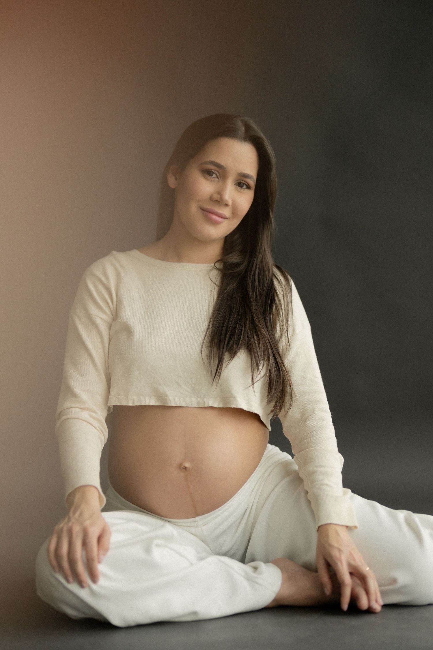 Maternity portrait of an expectant mother in white loungewear sitting cross-legged against a dark background.