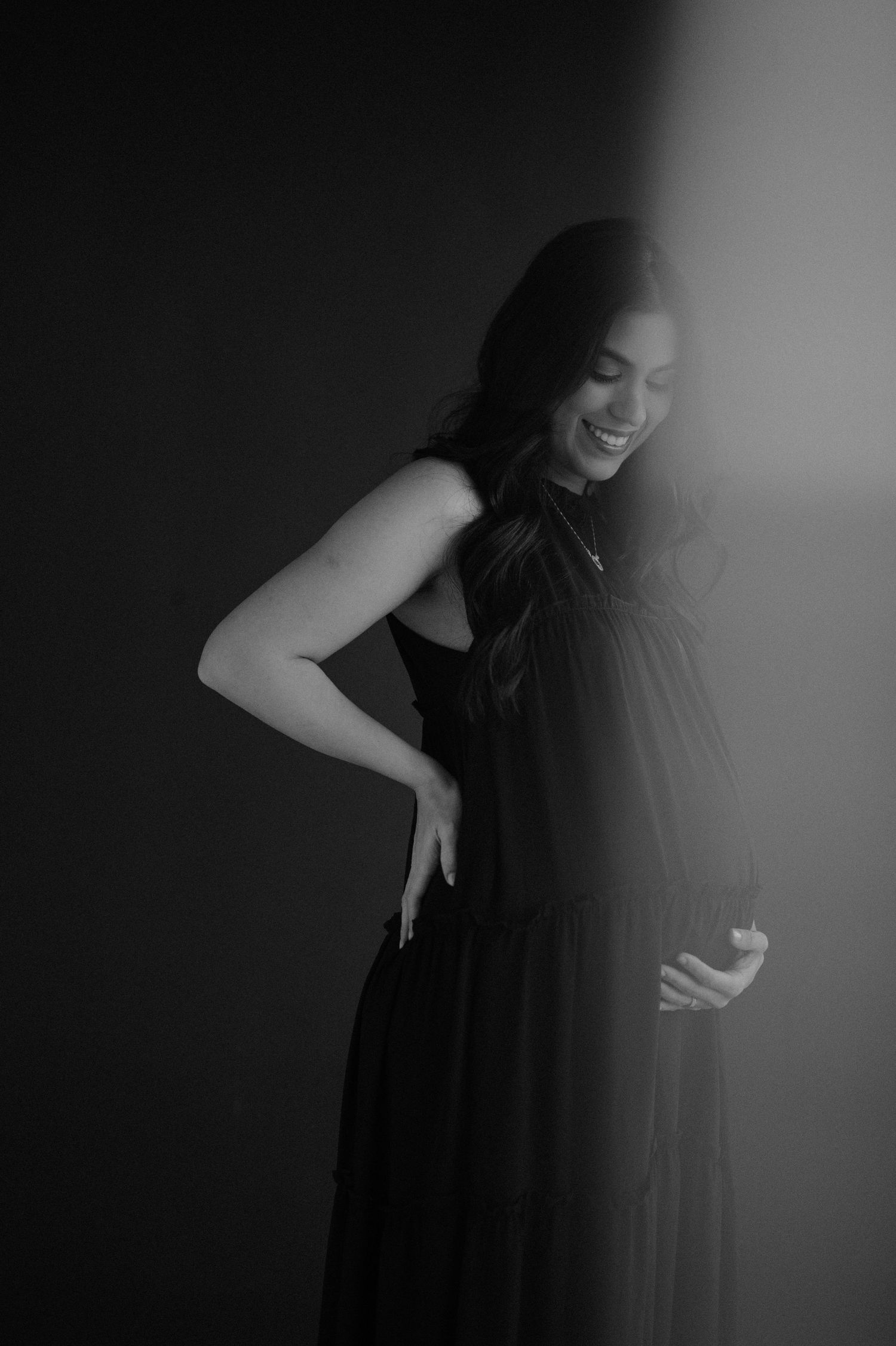 A woman in a black dress laughs joyfully against a dark background in this artistic black and white portrait.