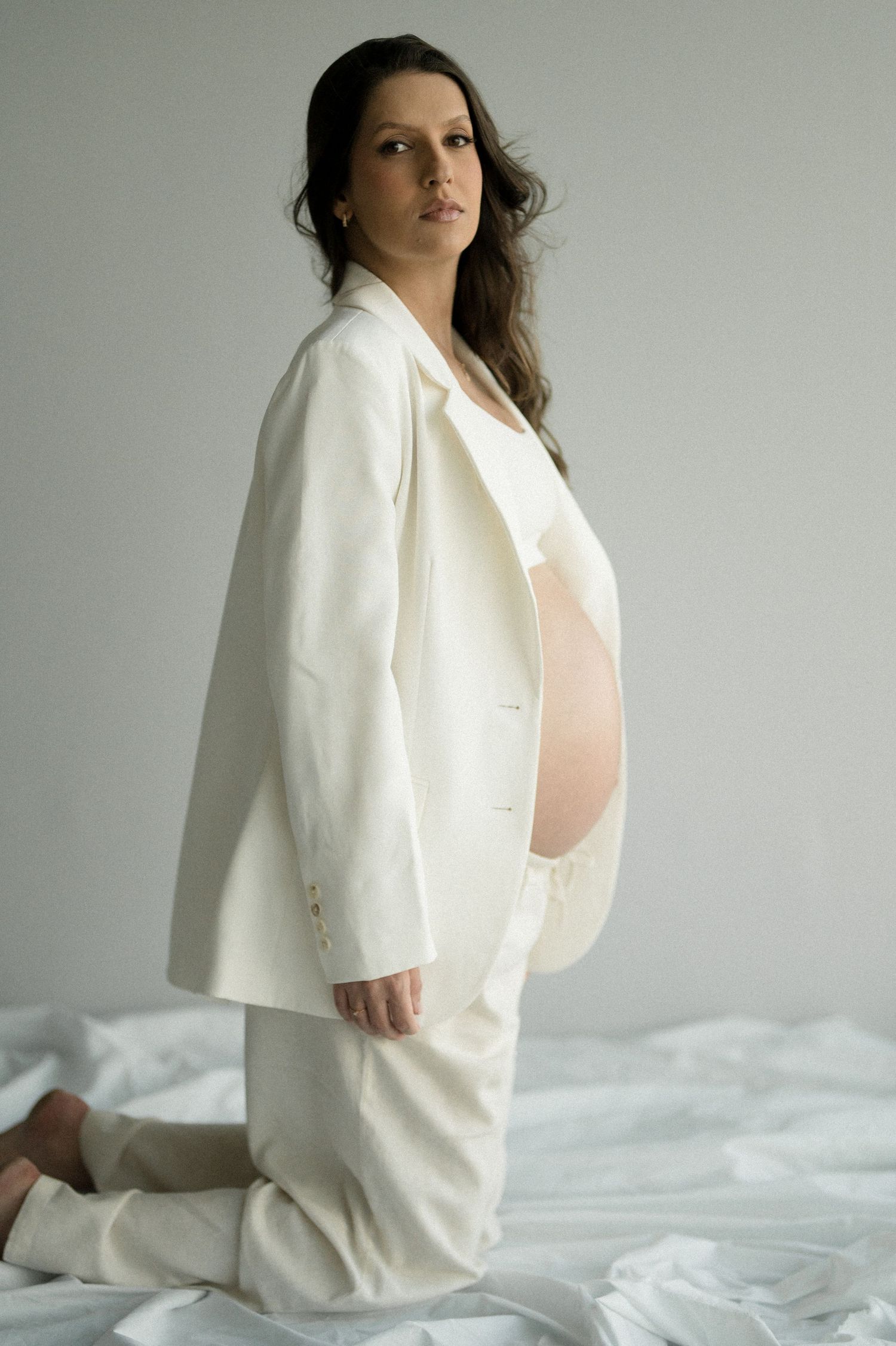 A person in a white blazer and pink top poses on white bedding in a minimalist setting.