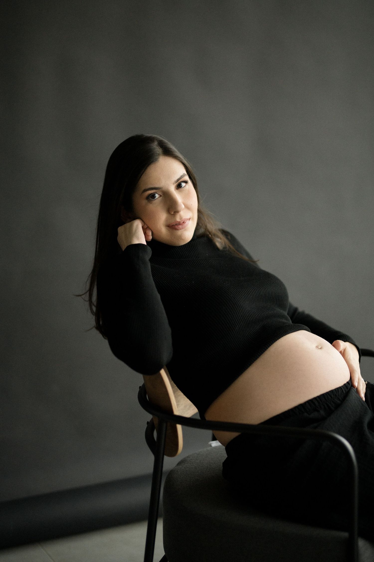 Artistic maternity photo featuring a pregnant woman in black attire seated on a chair against dark backdrop.