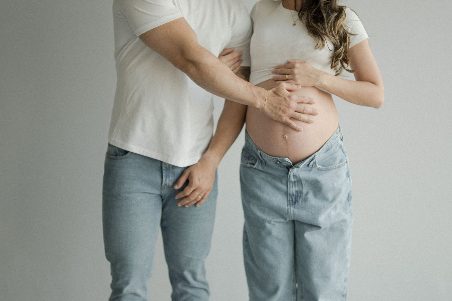 A couple in casual denim and white tops embrace while standing against a light gray background.