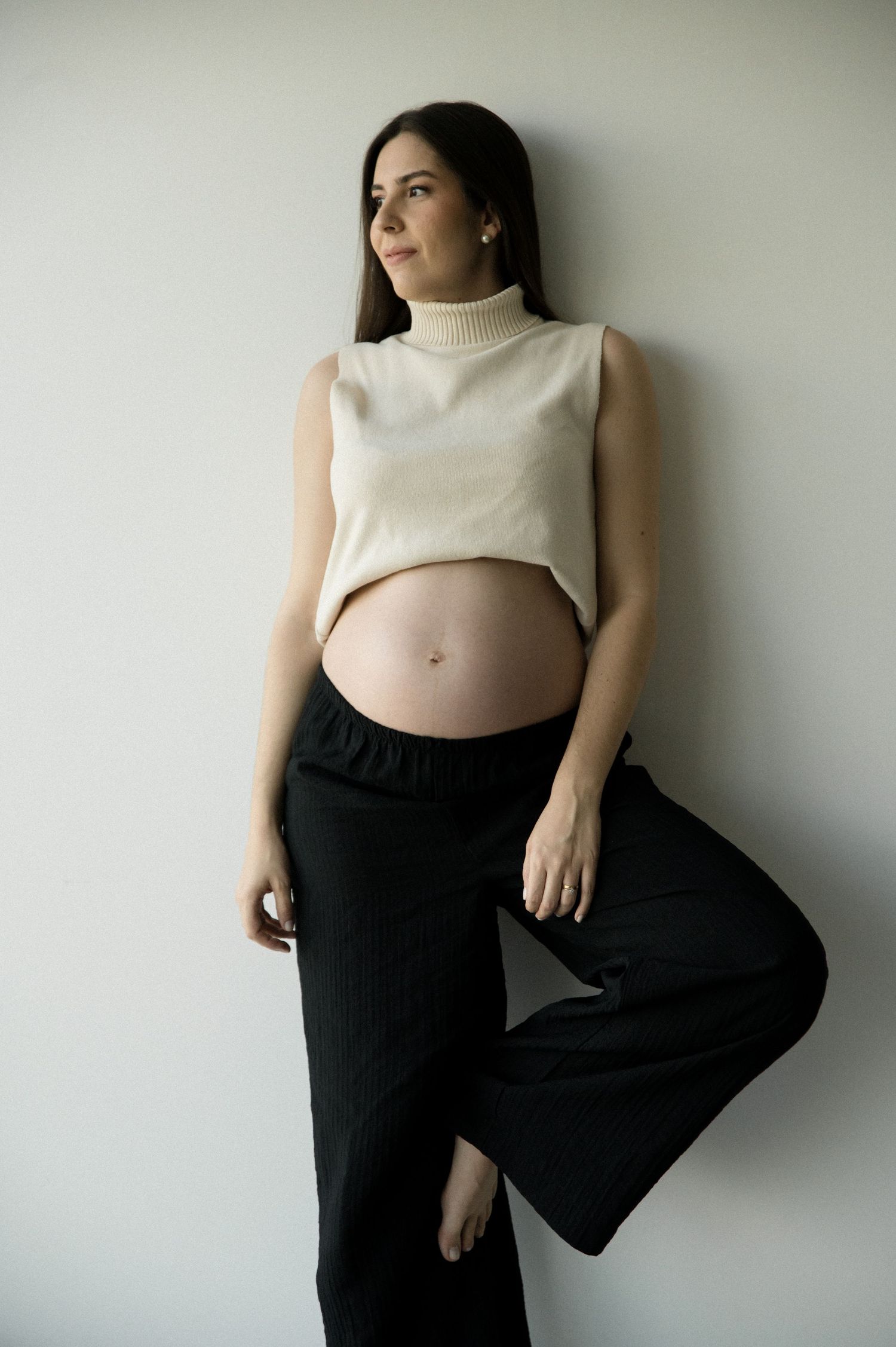 Modern maternity portrait showing an expecting mother in cream top and black pants against white wall.