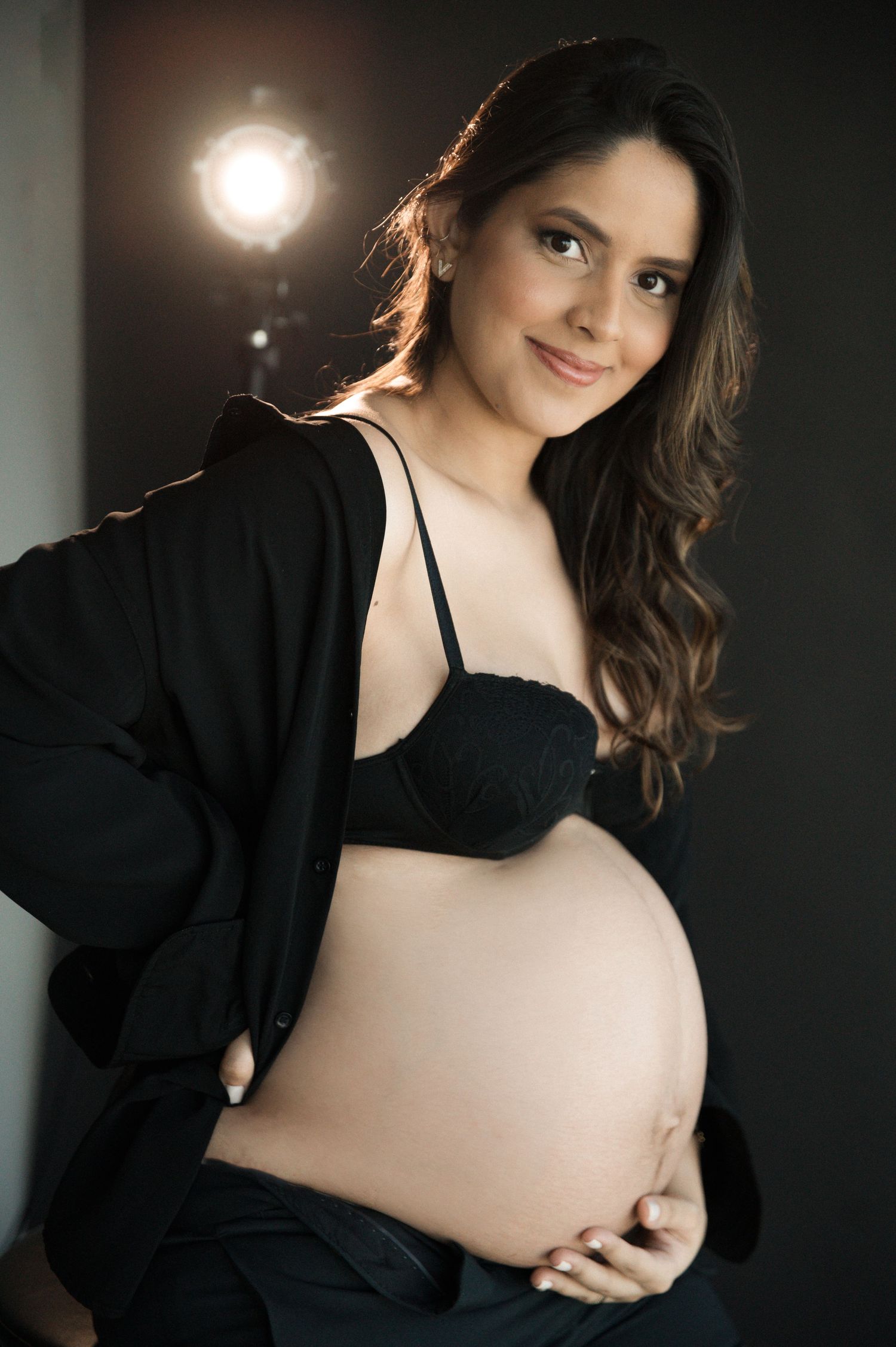 Pregnant woman in black bra and open jacket cradling baby bump, smiling softly in studio with backlight.