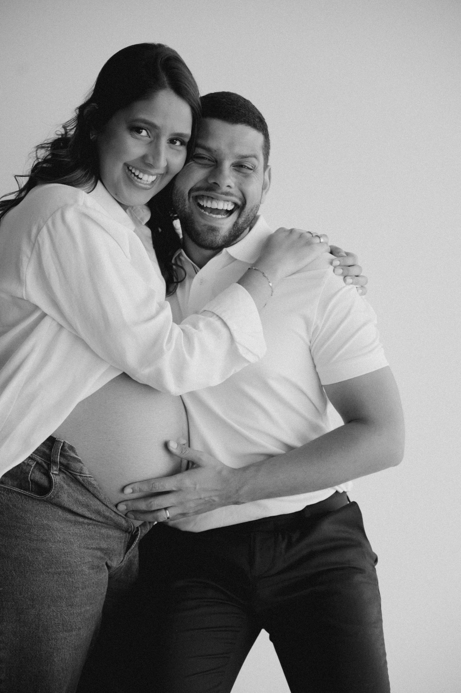 Black and white maternity photo of a smiling couple embracing, man tenderly touching pregnant woman's bare belly.