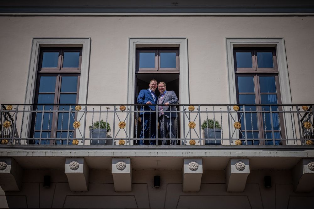 People stand on a balcony of a white building with three blue-framed windows and ornate architectural details.