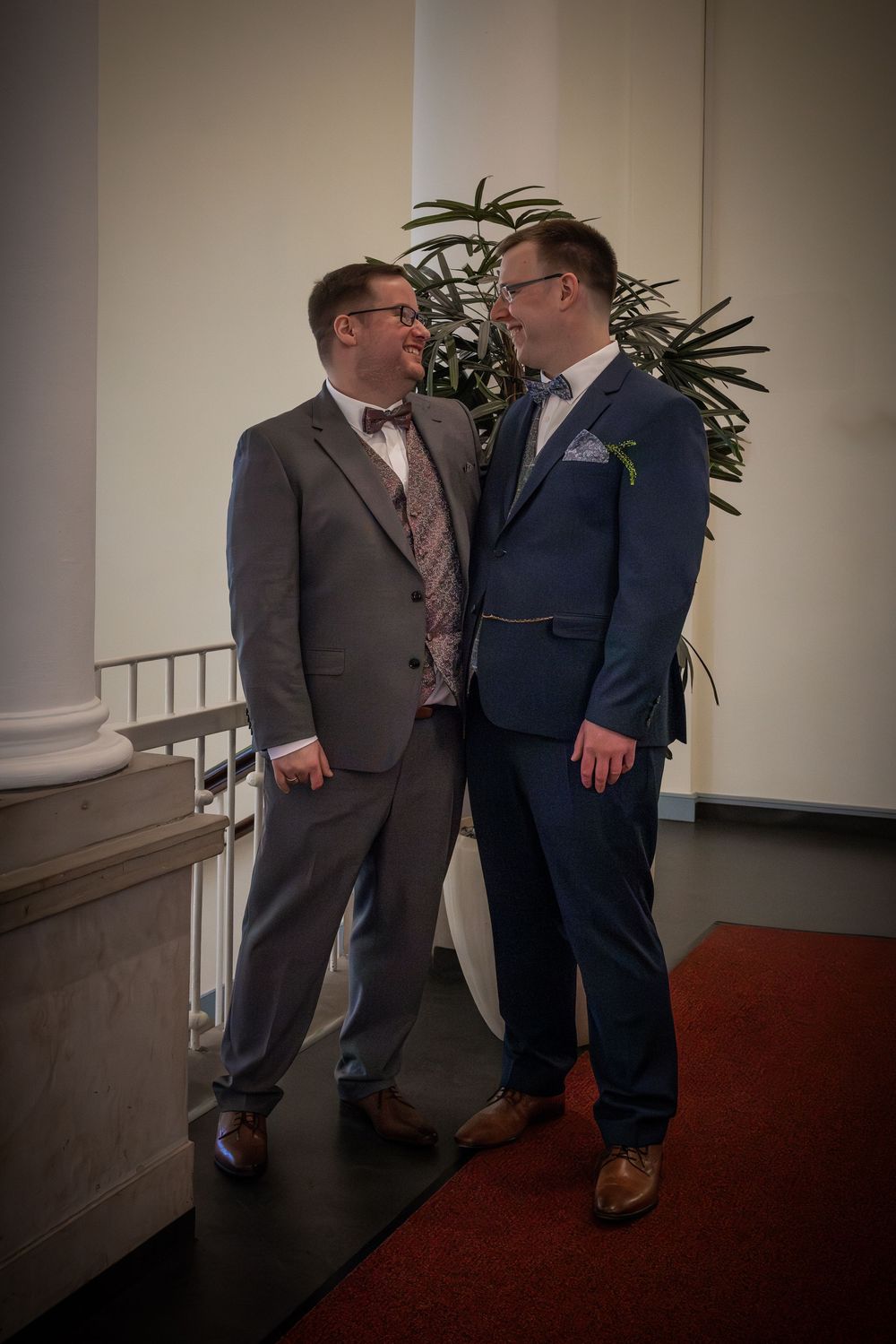 Two people in formal suits share a moment together at an indoor venue with white columns and potted plants in the background.