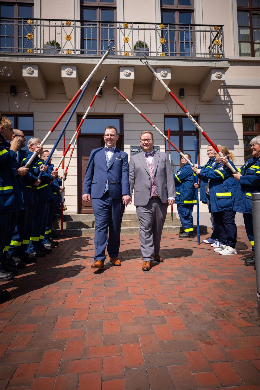 Two men in suits walk under an arch of crossed poles held by emergency response personnel on brick pavement.