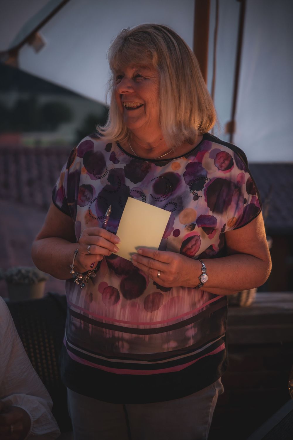 Person in purple floral top smiles while reading a letter or card in dim evening lighting.