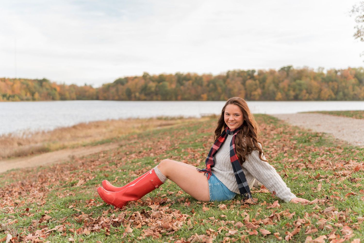 Someone in red rain boots sits on autumn leaves by a lake with fall foliage in the background.