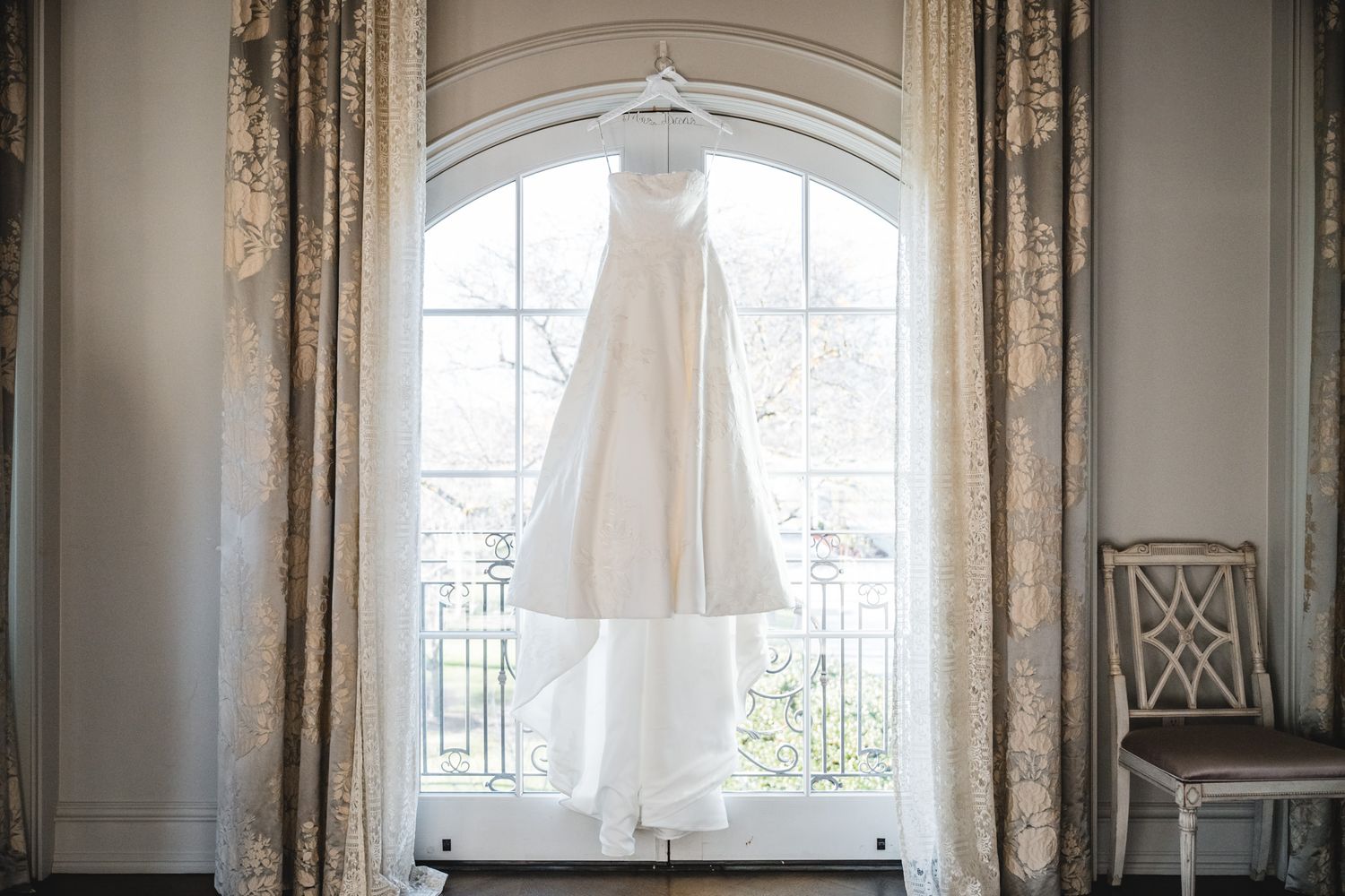 White wedding dress hangs elegantly in front of a large arched window with natural light.