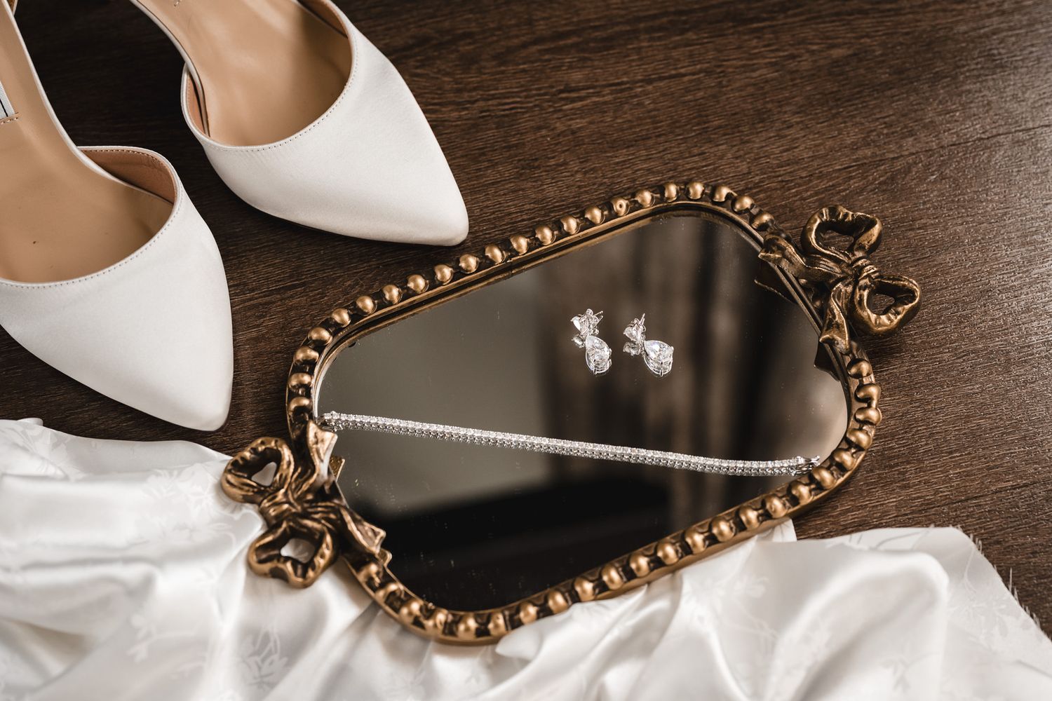 Wedding accessories including white shoes, jewelry and ornate mirror on wooden surface.