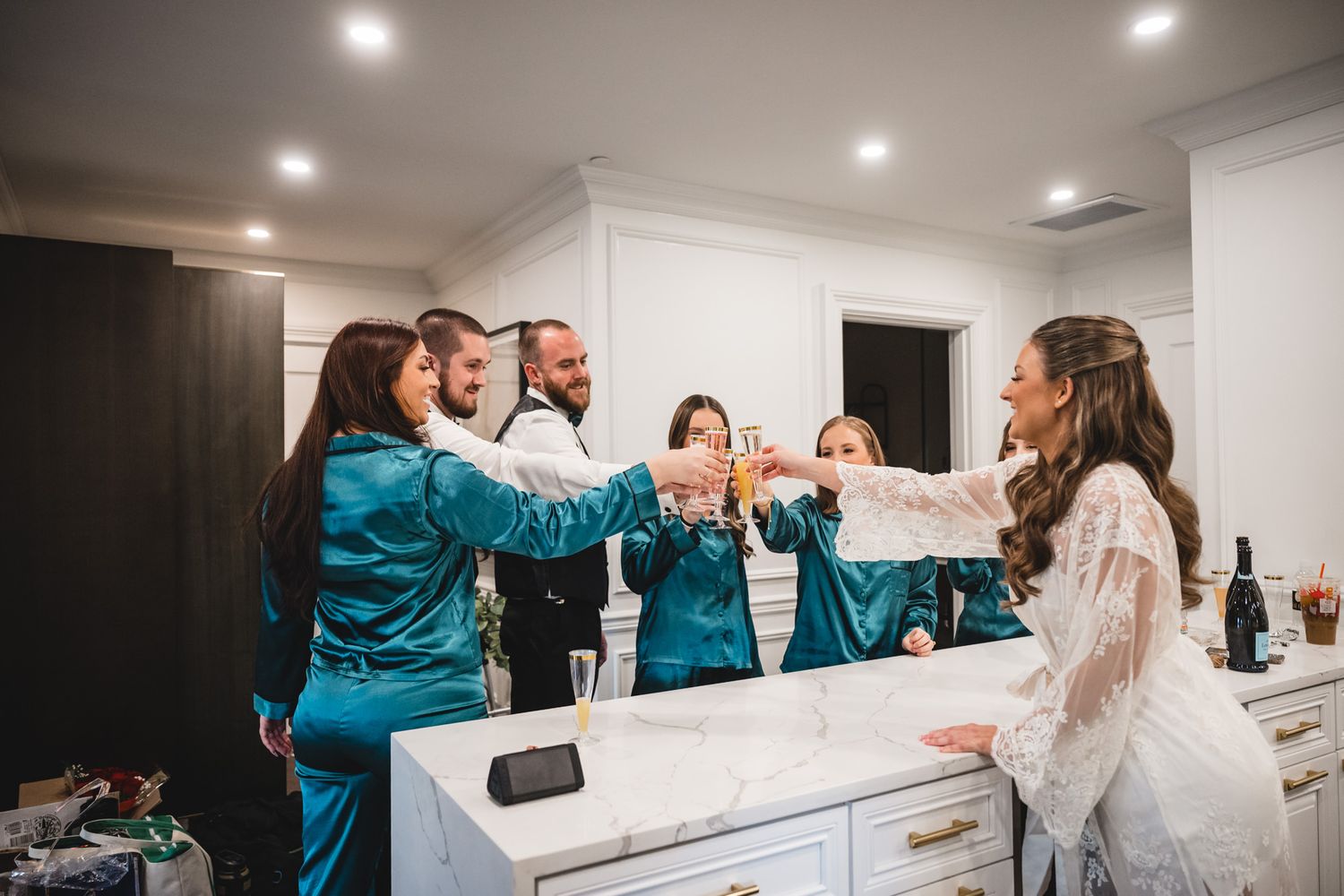 Wedding party members laugh together during preparation in a modern white bathroom.
