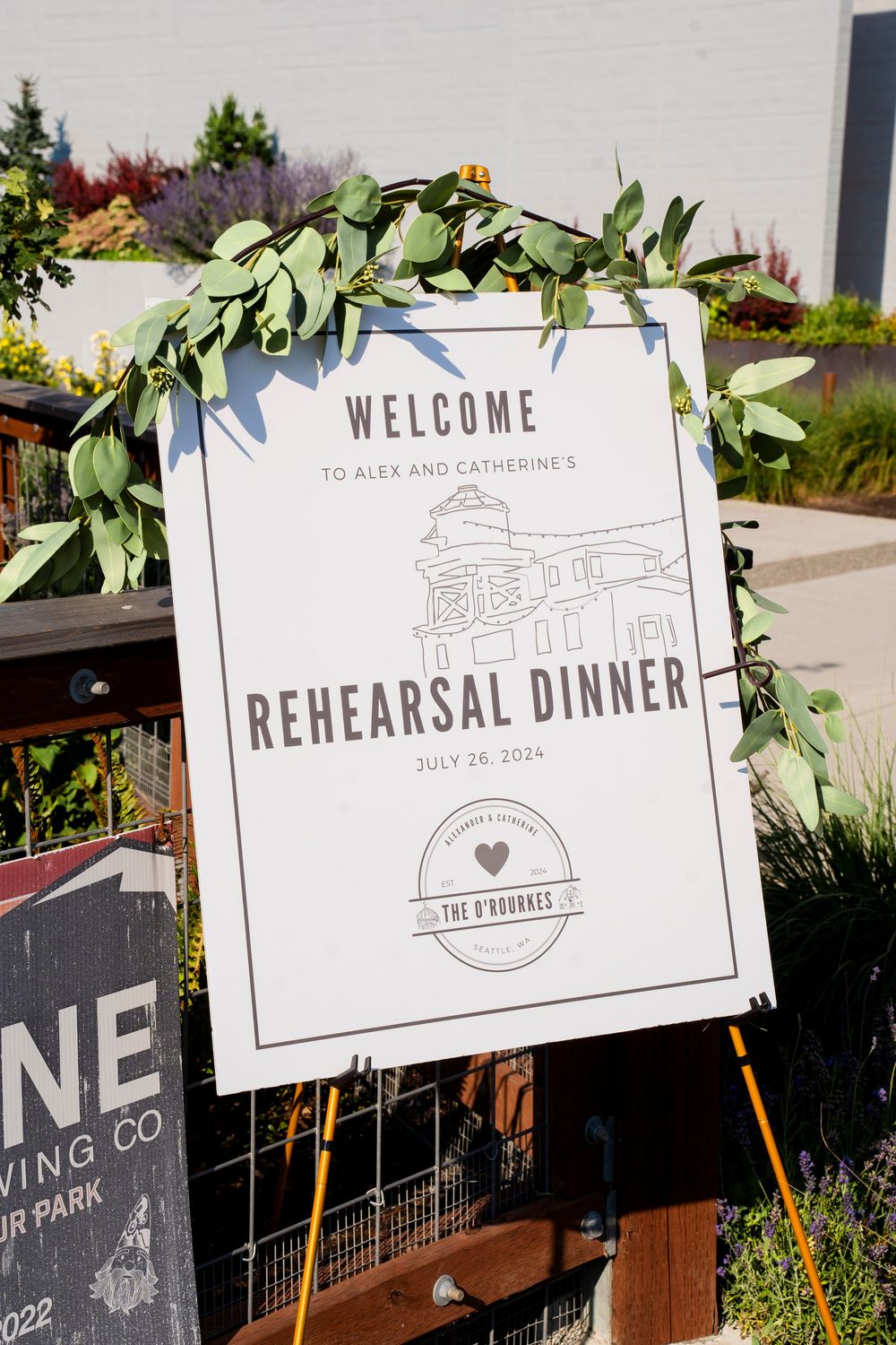 A wedding rehearsal dinner welcome sign decorated with eucalyptus leaves on an easel stand outdoors.