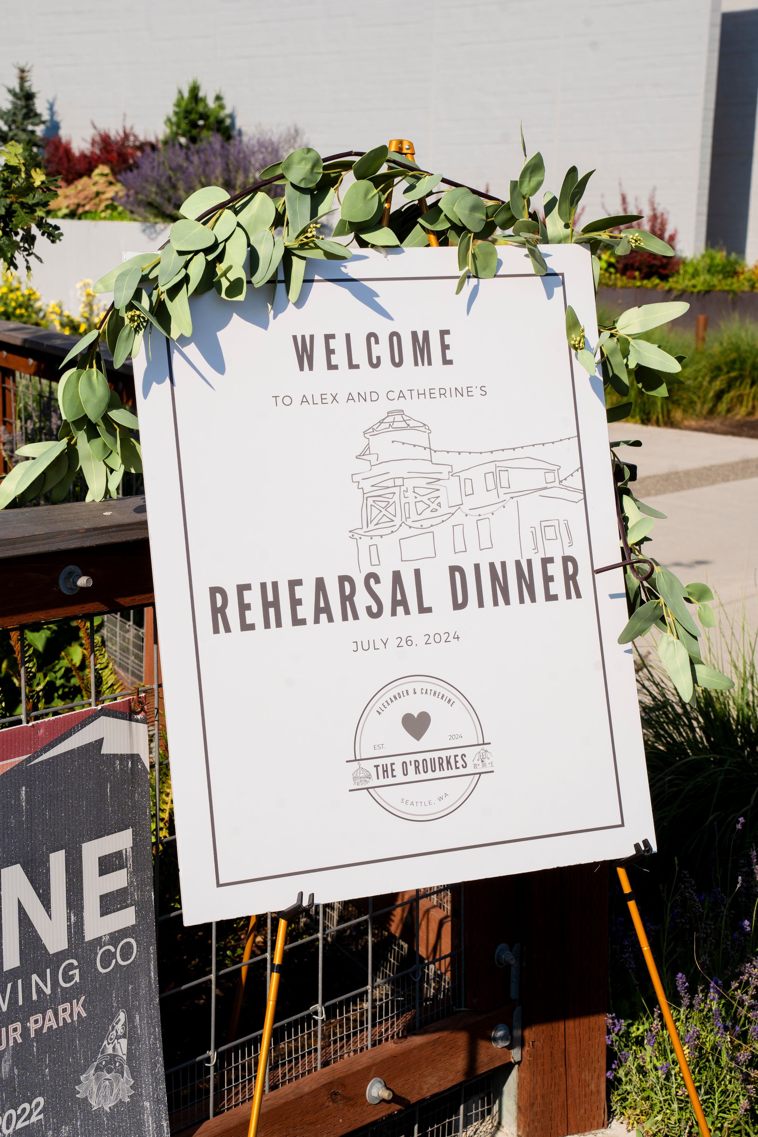 A wedding rehearsal dinner welcome sign decorated with eucalyptus leaves on an easel stand outdoors.