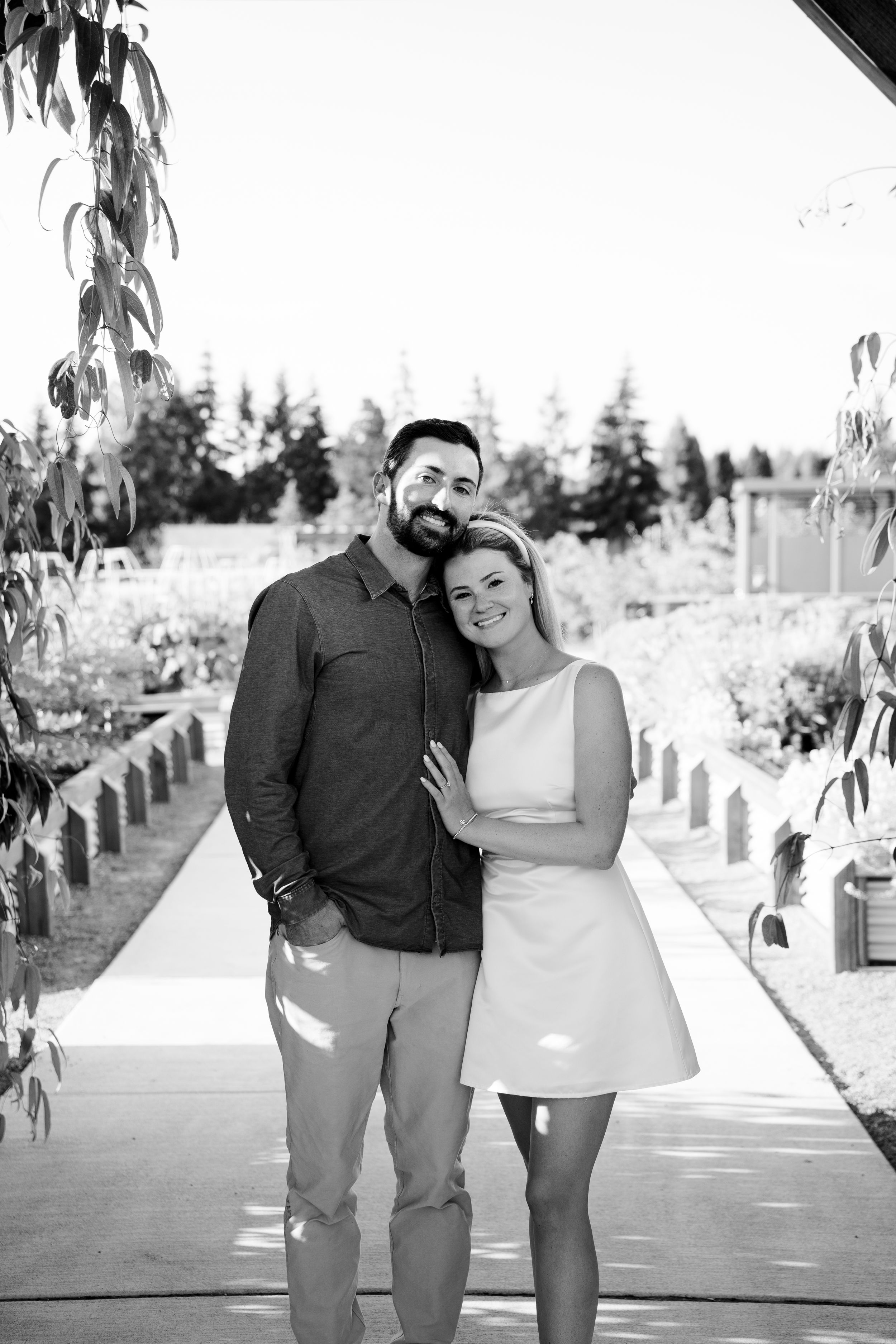 A black and white photo of a couple standing together on a garden pathway surrounded by greenery.