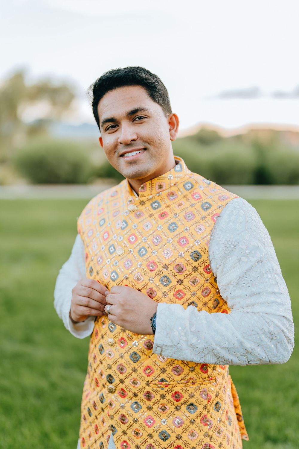 A groom in a yellow patterned vest and white kurta smiles joyfully during his wedding celebration.