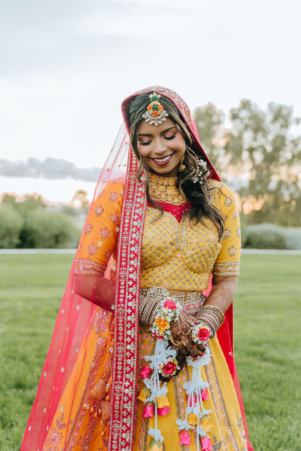 Bride in vibrant yellow and red lehenga with traditional jewelry in outdoor garden setting.