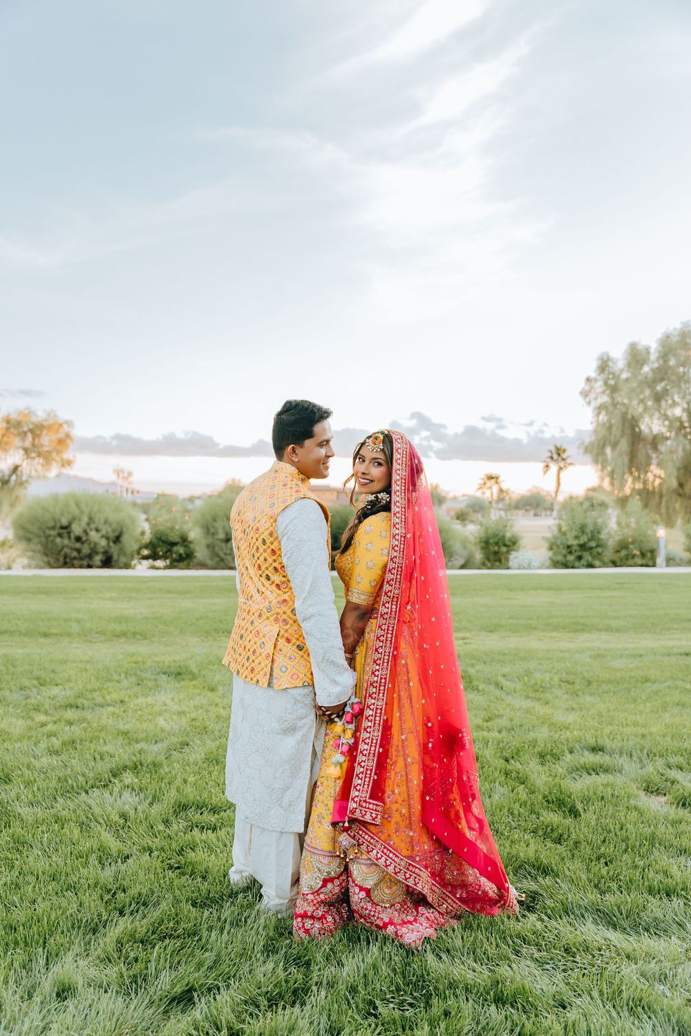 Wedding couple in traditional South Asian attire poses in outdoor garden setting.