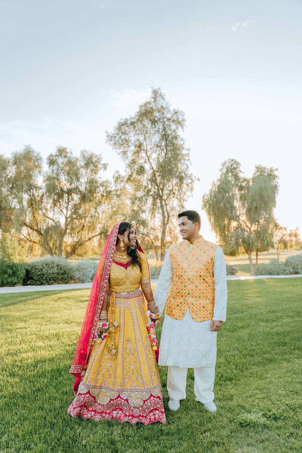 Wedding couple in traditional yellow and white Indian attire poses together in a sunlit garden setting.