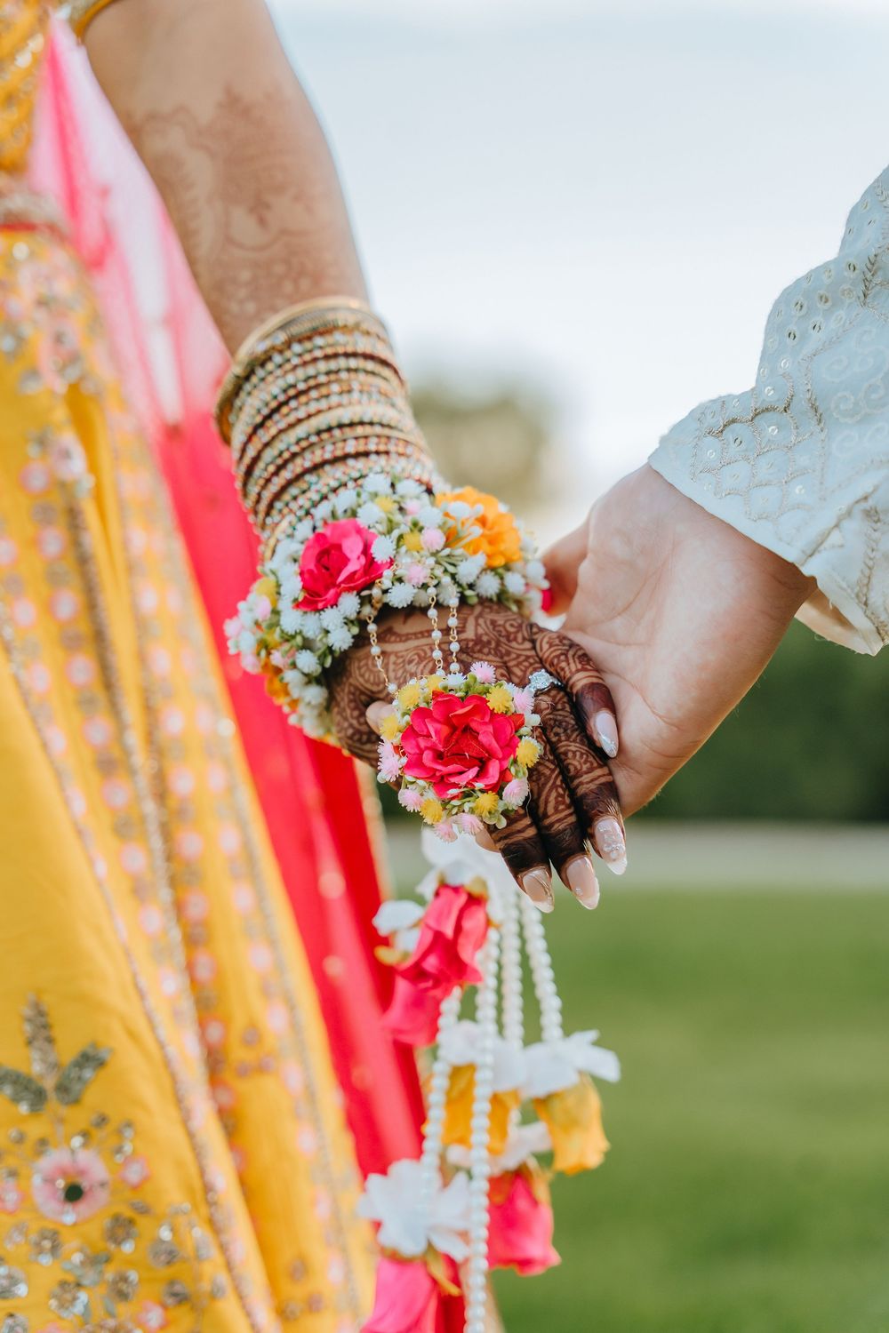 Close-up view of hands adorned with bangles holding colorful floral bouquets against a vibrant yellow and pink fabric backdrop.
