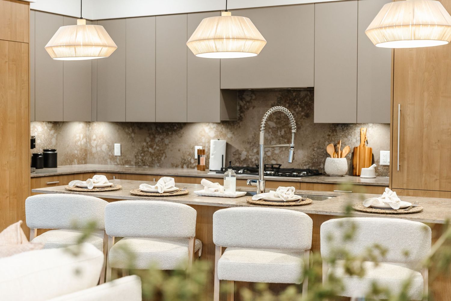 Modern kitchen interior with pendant lights, marble backsplash, and white upholstered bar stools at wooden counter.