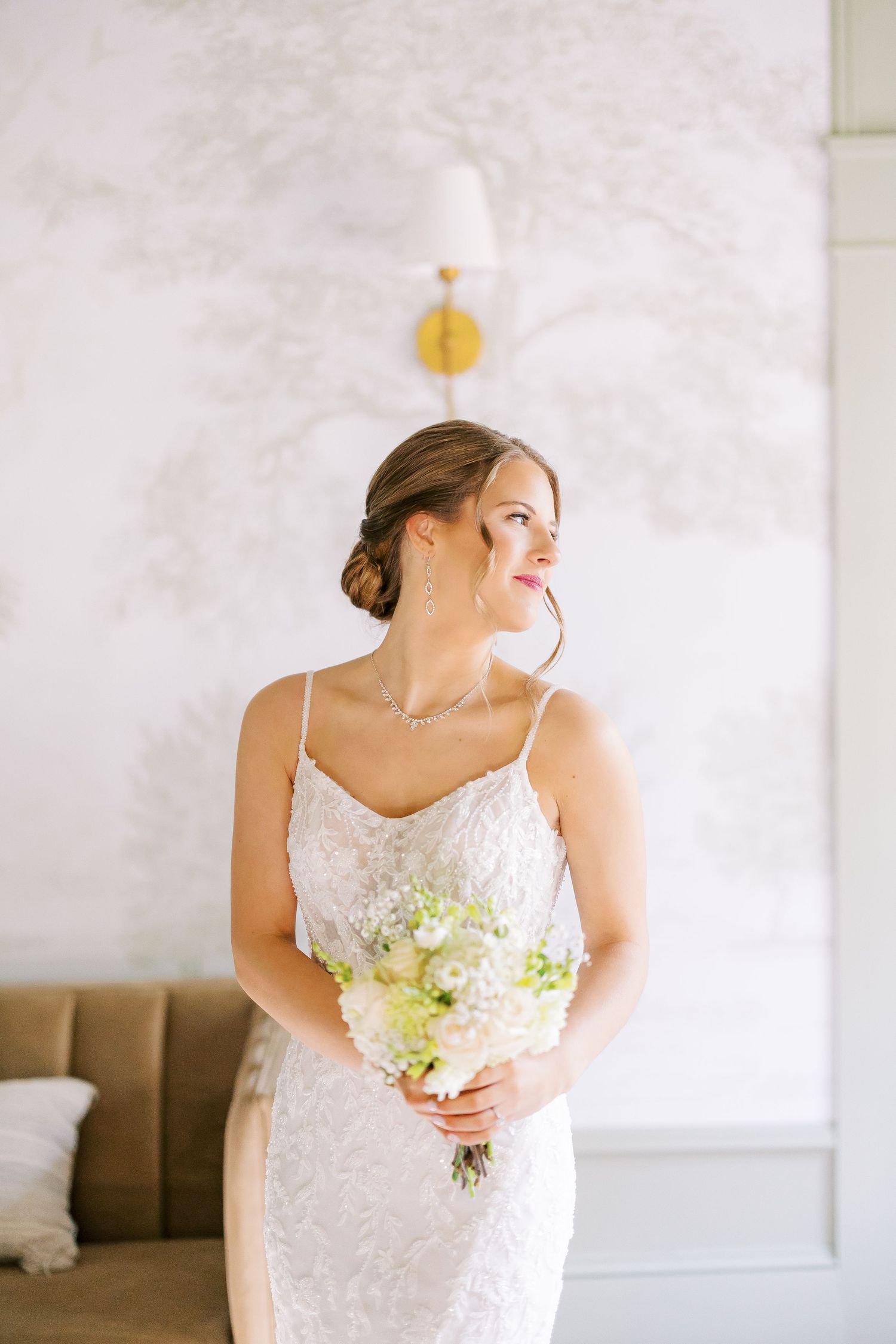 A bride in a delicate lace wedding dress holds a white and green bouquet while posing in a bright interior space.