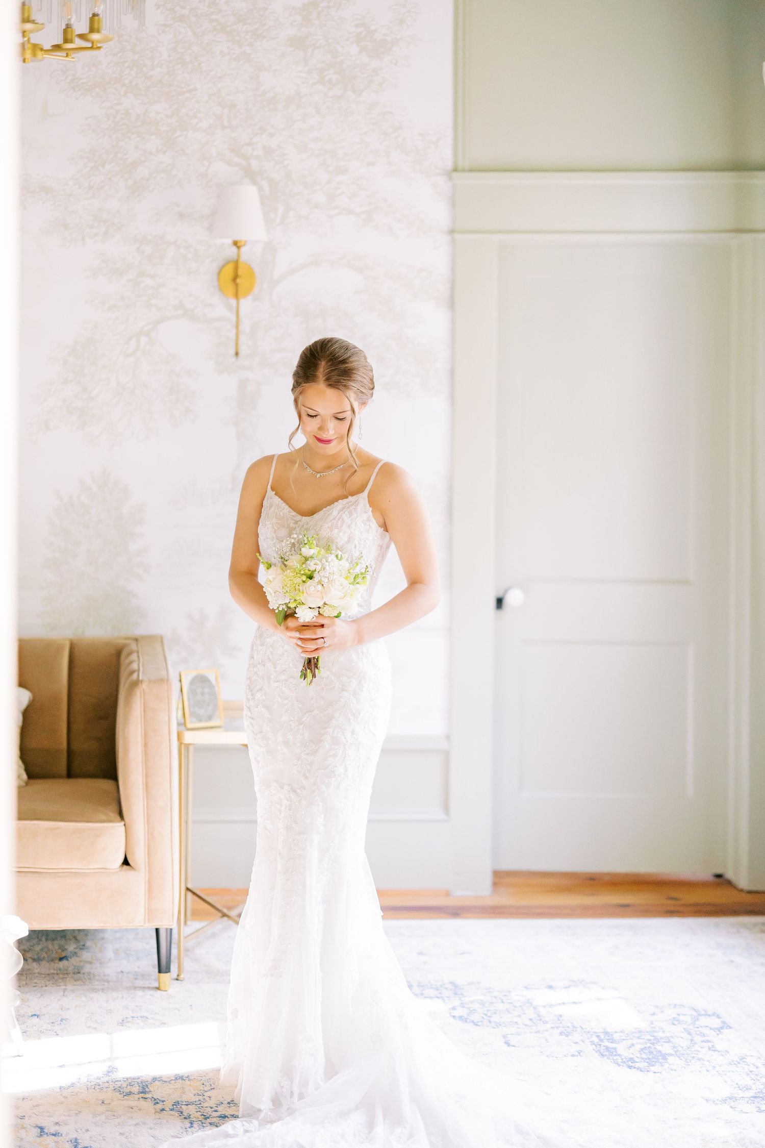 A bride in an elegant white lace mermaid wedding gown holds a white bouquet while standing in a sunlit room.