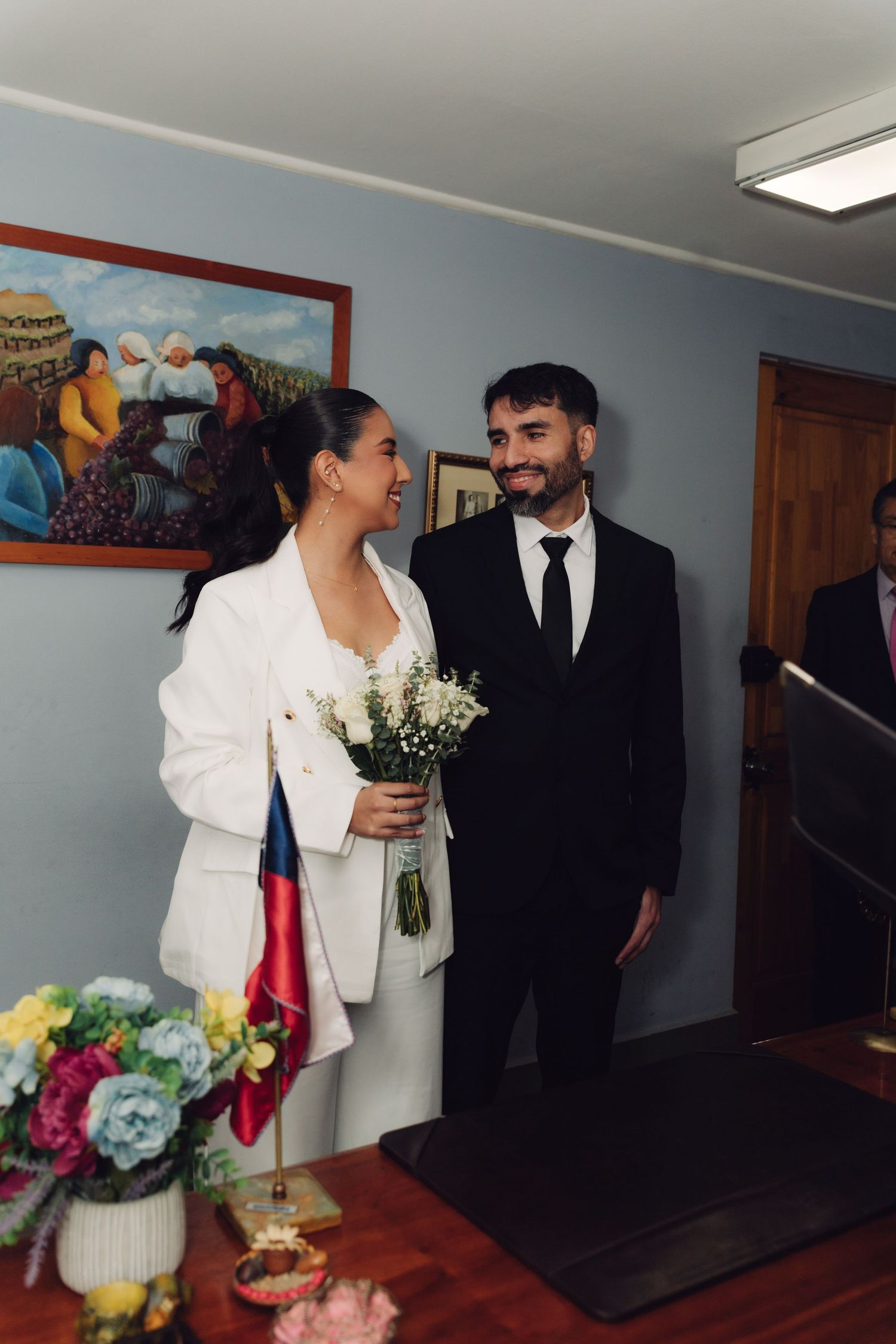 Bride in white suit and groom in black suit at civil wedding ceremony with Chilean flag.
