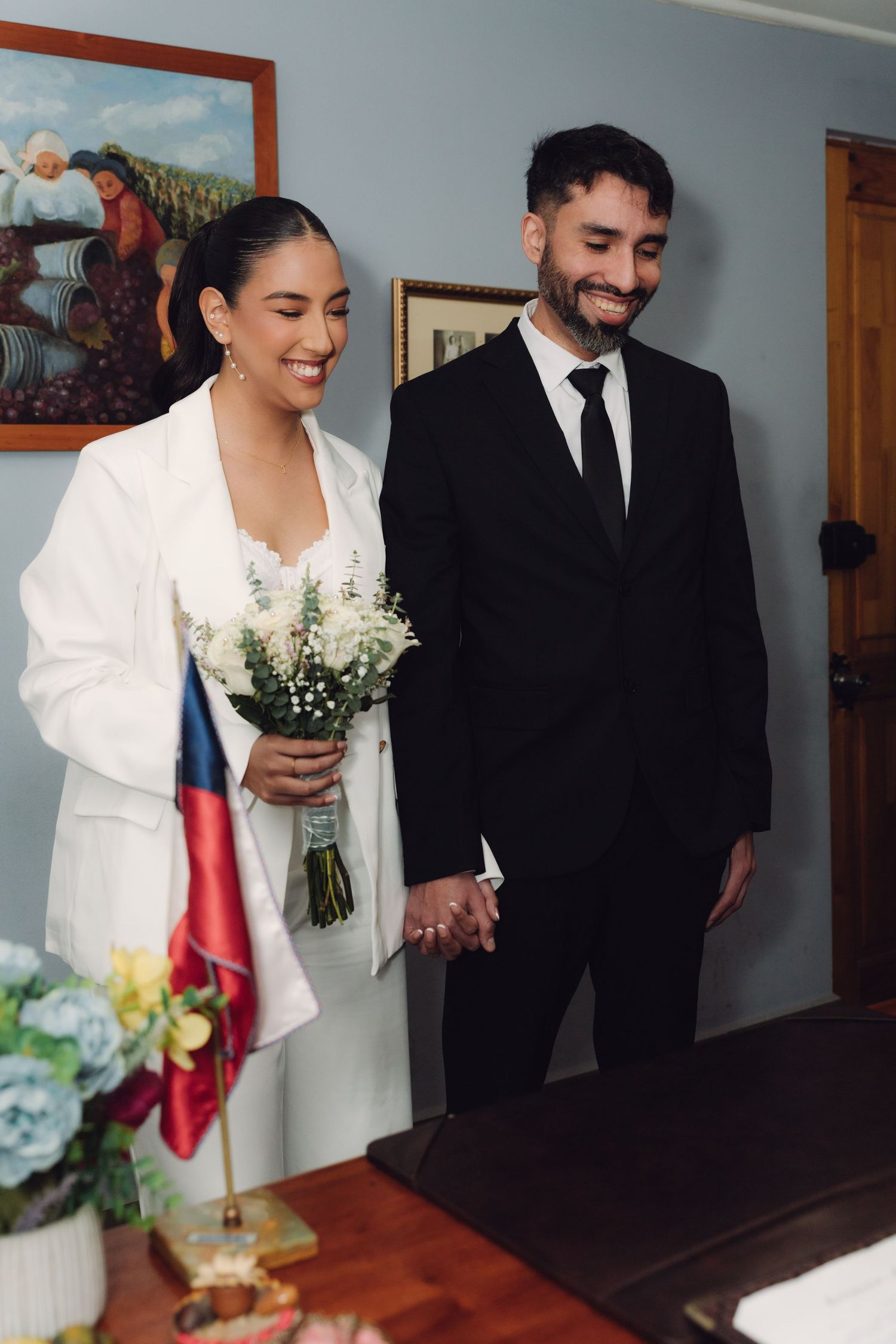 Smiling newlyweds holding hands at civil registry office with Chilean flag in background.