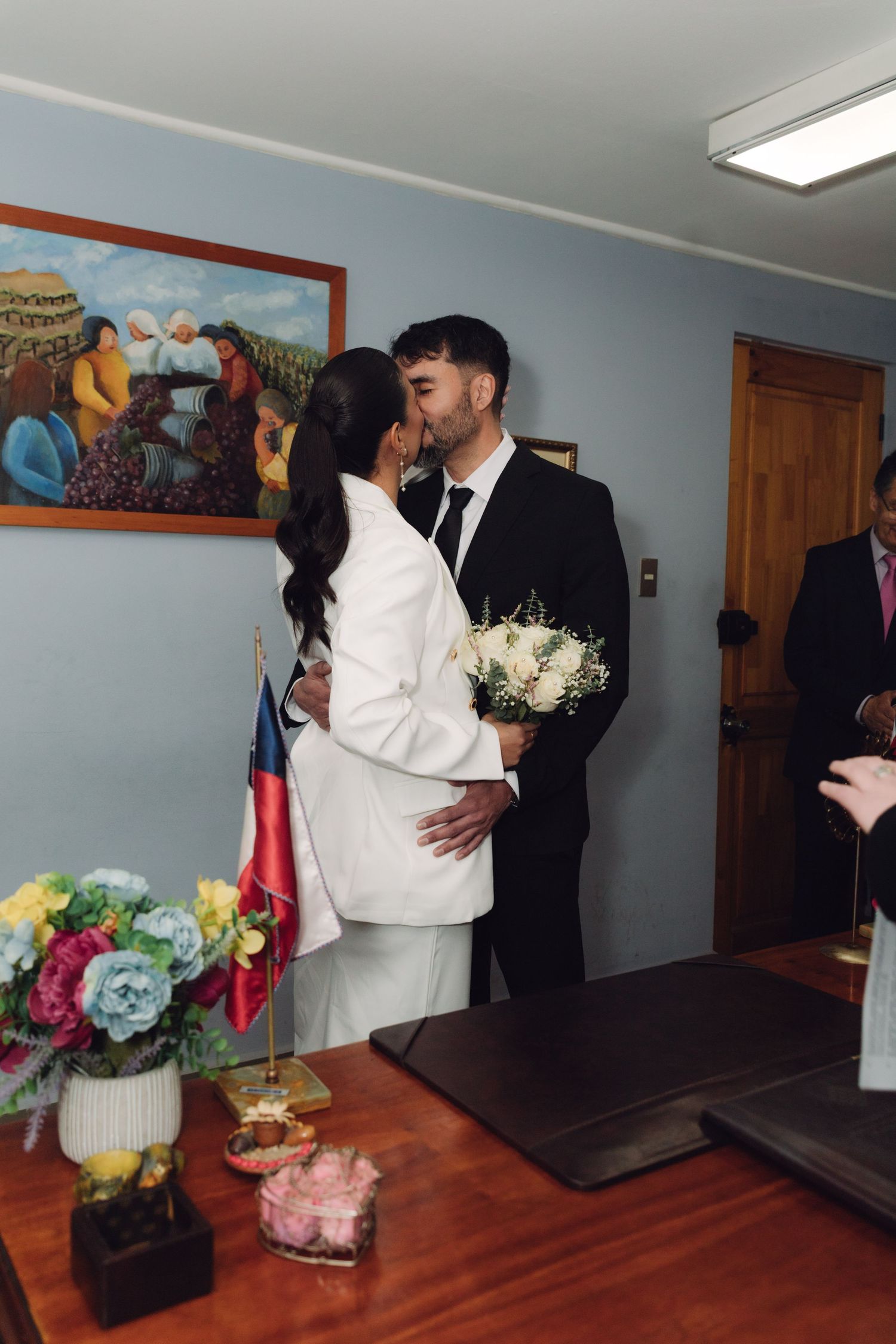 Bride and groom sharing first kiss at civil wedding ceremony with Chilean flag present.
